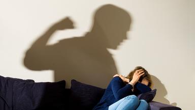 London April 2nd, 2021, A woman holds her arms protectively in front of her face while she sits on the sofa and the shadow of a man can be seen with a clenched fist.