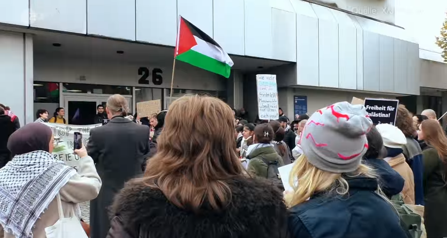 Mehrere Menschen versammeln sich zu einer pro-palästinensischen Demonstration vor einer Universität. Sie tragen Plakate und die palästinensische Nationalflagge.