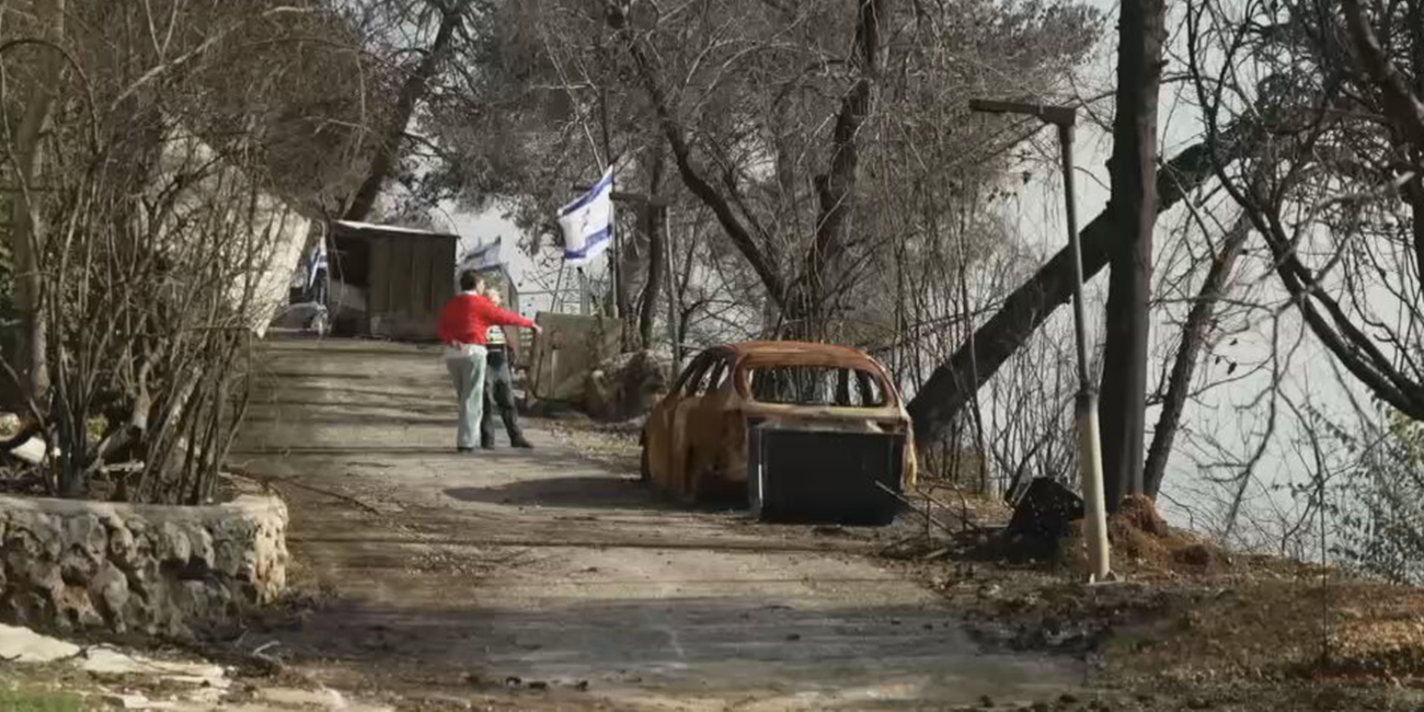 Es ist der zerstörte Kibbuz Menara zu erkennen, mit verbrannten Autos und Israel-Flaggen im Hintergrund.