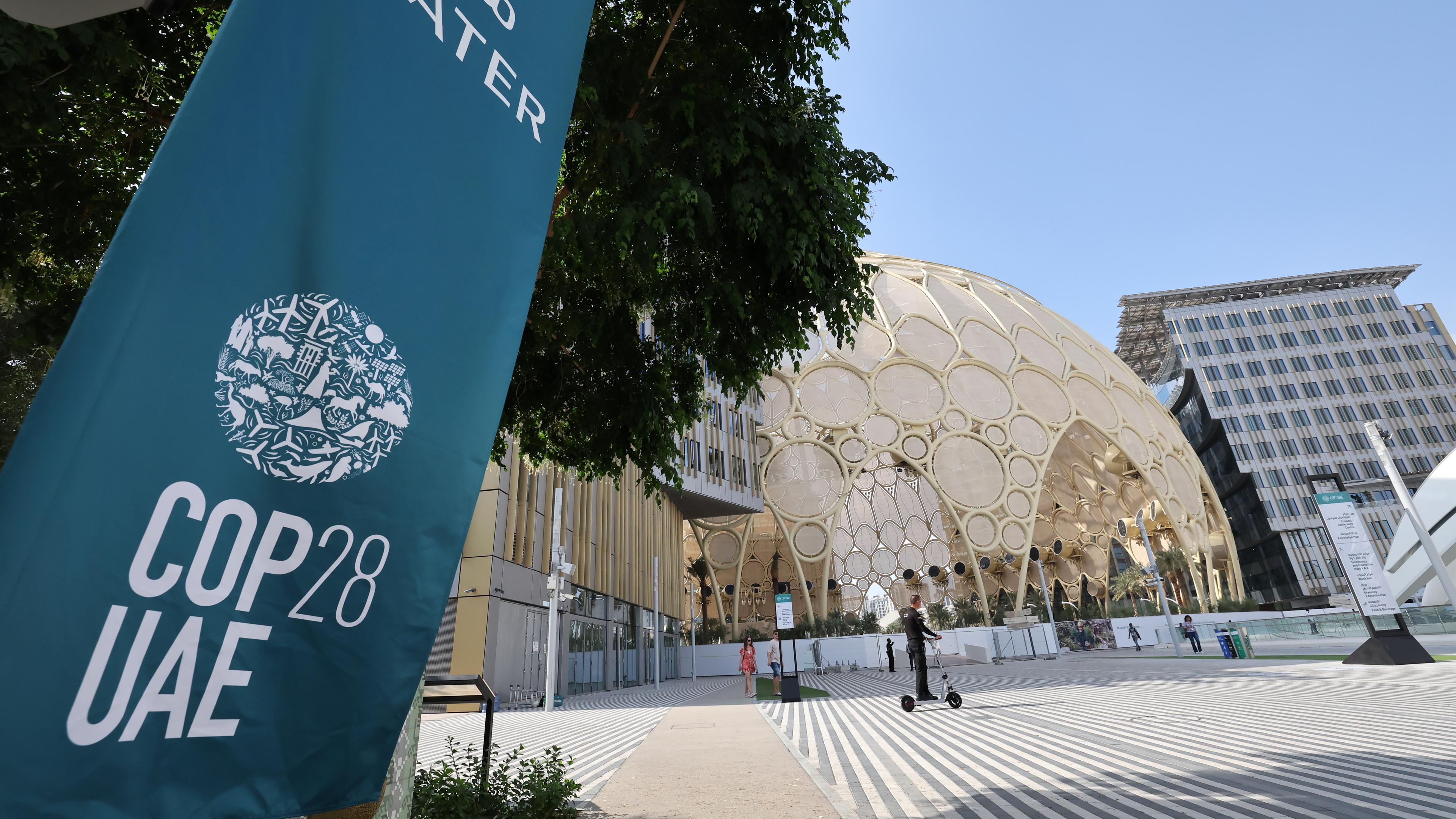 Logo COP28 auf einem Plakat, daneben ein Blick auf ein Gebäude