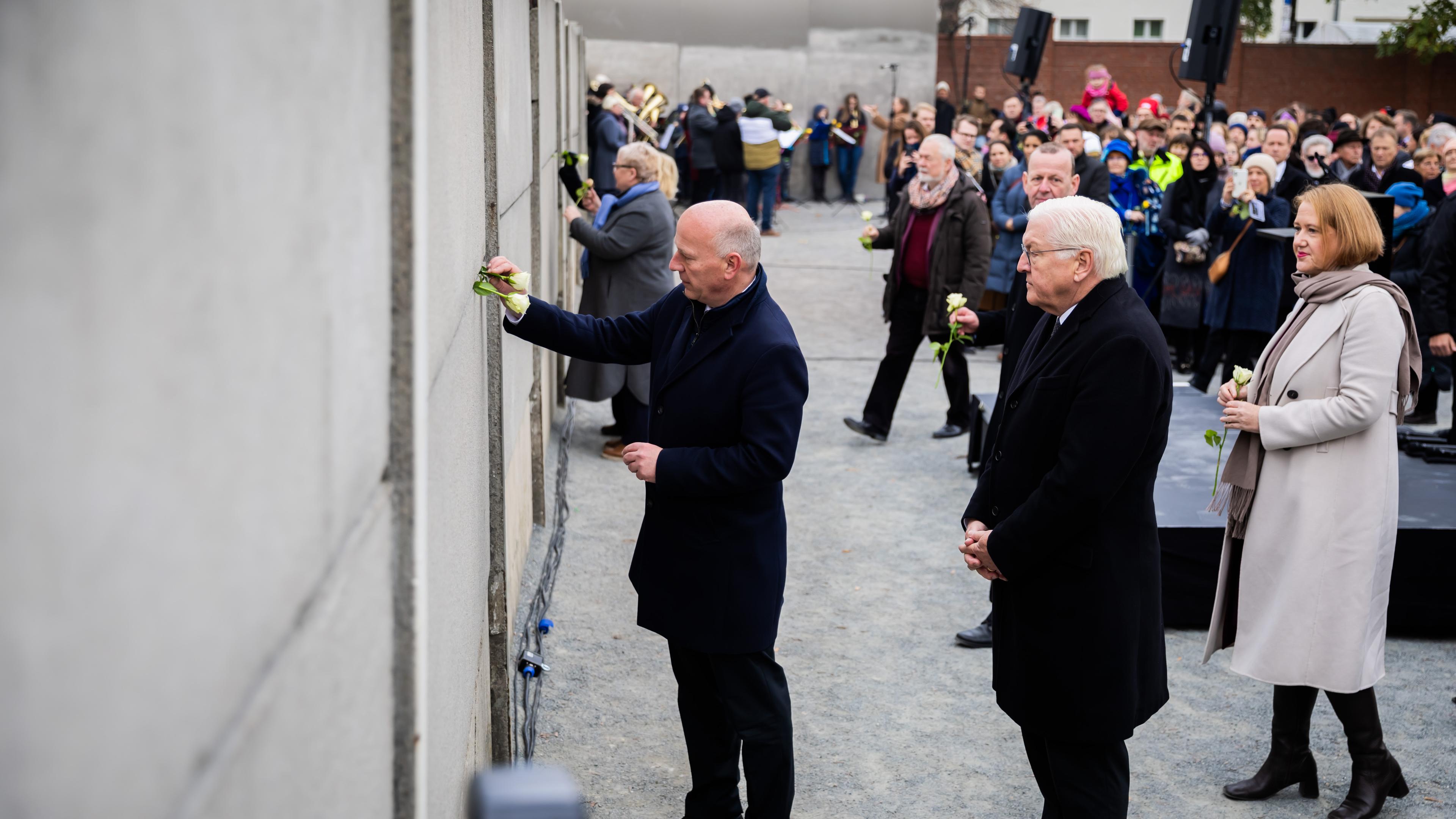 Kai Wegner (CDU, l), Regierender Bürgermeister von Berlin, steckt neben Bundespräsident Frank-Walter Steinmeier und Lisa Paus (Bündnis 90/Die Grünen), Bundesministerin für Familie, Senioren, Frauen und Jugend, bei einer zentralen Gedenkveranstaltung anlässlich von 35 Jahre friedliche Revolution und Mauerfall in der Gedenkstätte Berliner Mauer eine Rose in die Hinterlandmauer. 