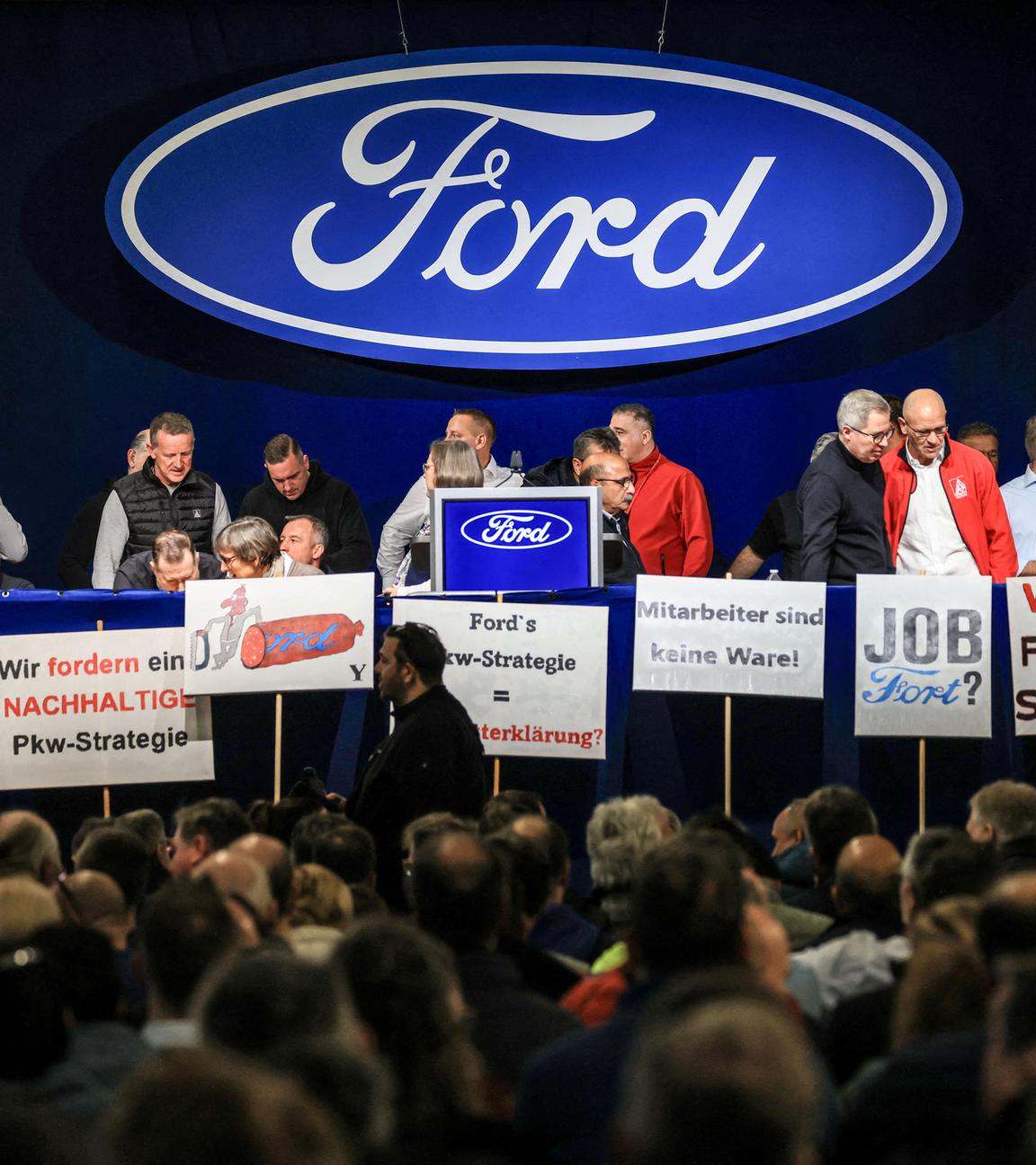 Mitarbeiter, Gewerkschaftler stehen auf einer Bühne mit Schildern, dahinter ein Ford Logo