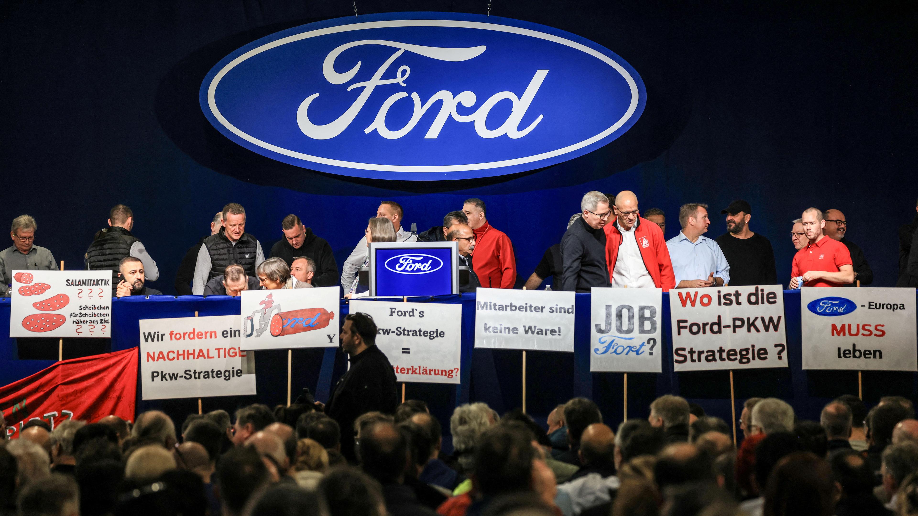 Mitarbeiter, Gewerkschaftler stehen auf einer Bühne mit Schildern, dahinter ein Ford Logo