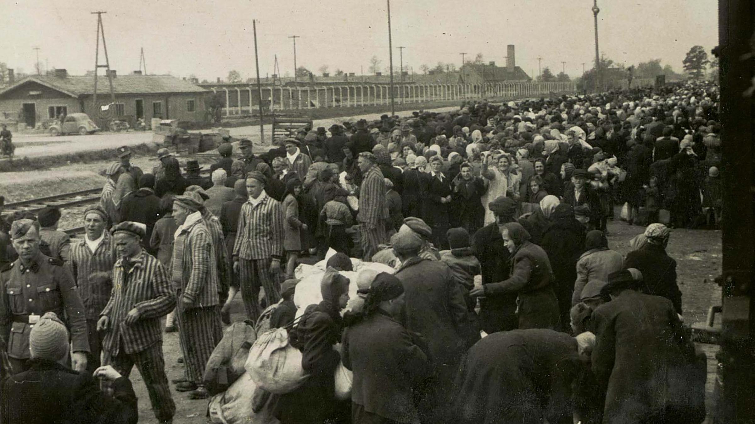 KZ Auschwitz-Birkenau - Die Menschen müssen nun schließlich ihr Gepäck endgültig ablegen und schauen besorgt auf den Bündelhaufen. 