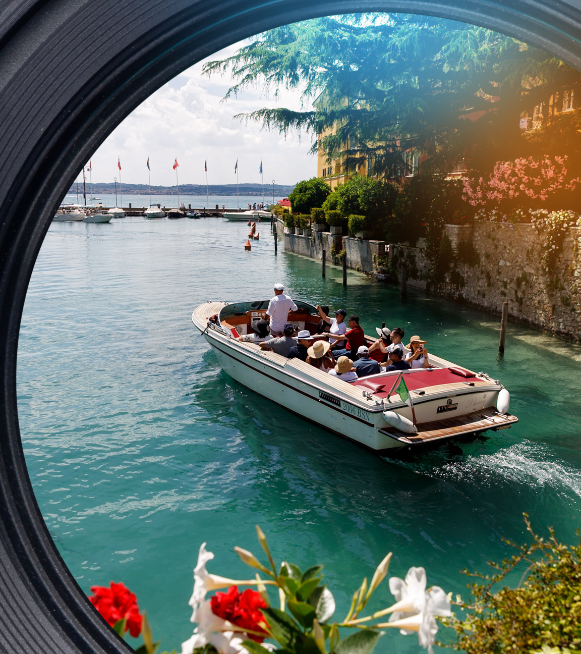 Ein Touristenboot, ein Wassertaxi, fährt an einer Mauer entlang auf dem Gardasee. Im Vordergrund blühen Blumen, im Hintergrund liegen weitere Schiffe.