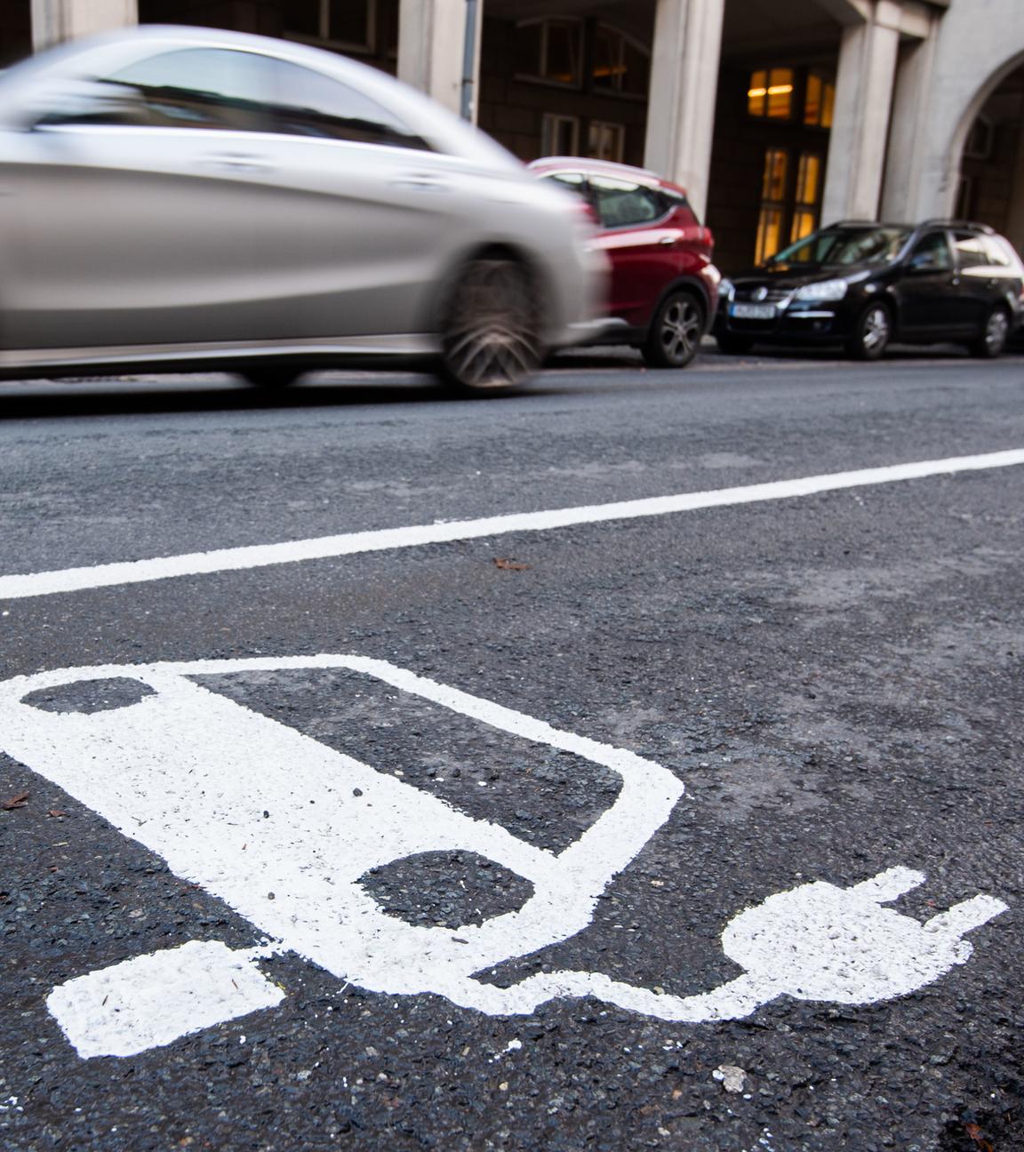 Ein symbol eines Elektroautos markiert einen Parkplatz neben einer E-Ladesaeule, während ein Auto vorbeifährt.