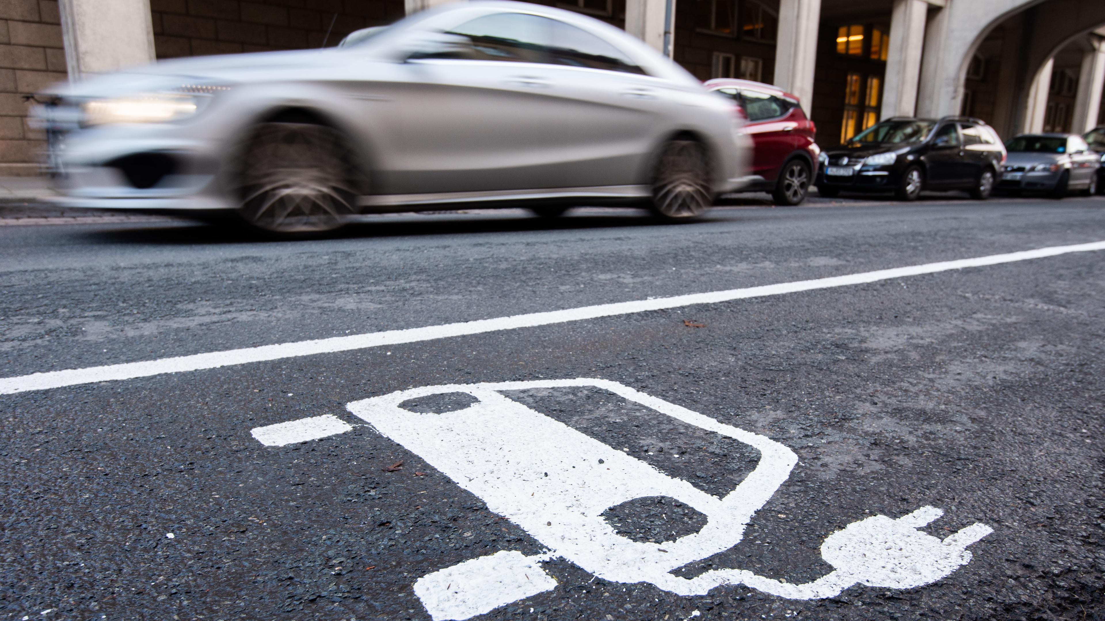 Ein symbol eines Elektroautos markiert einen Parkplatz neben einer E-Ladesaeule, während ein Auto vorbeifährt.