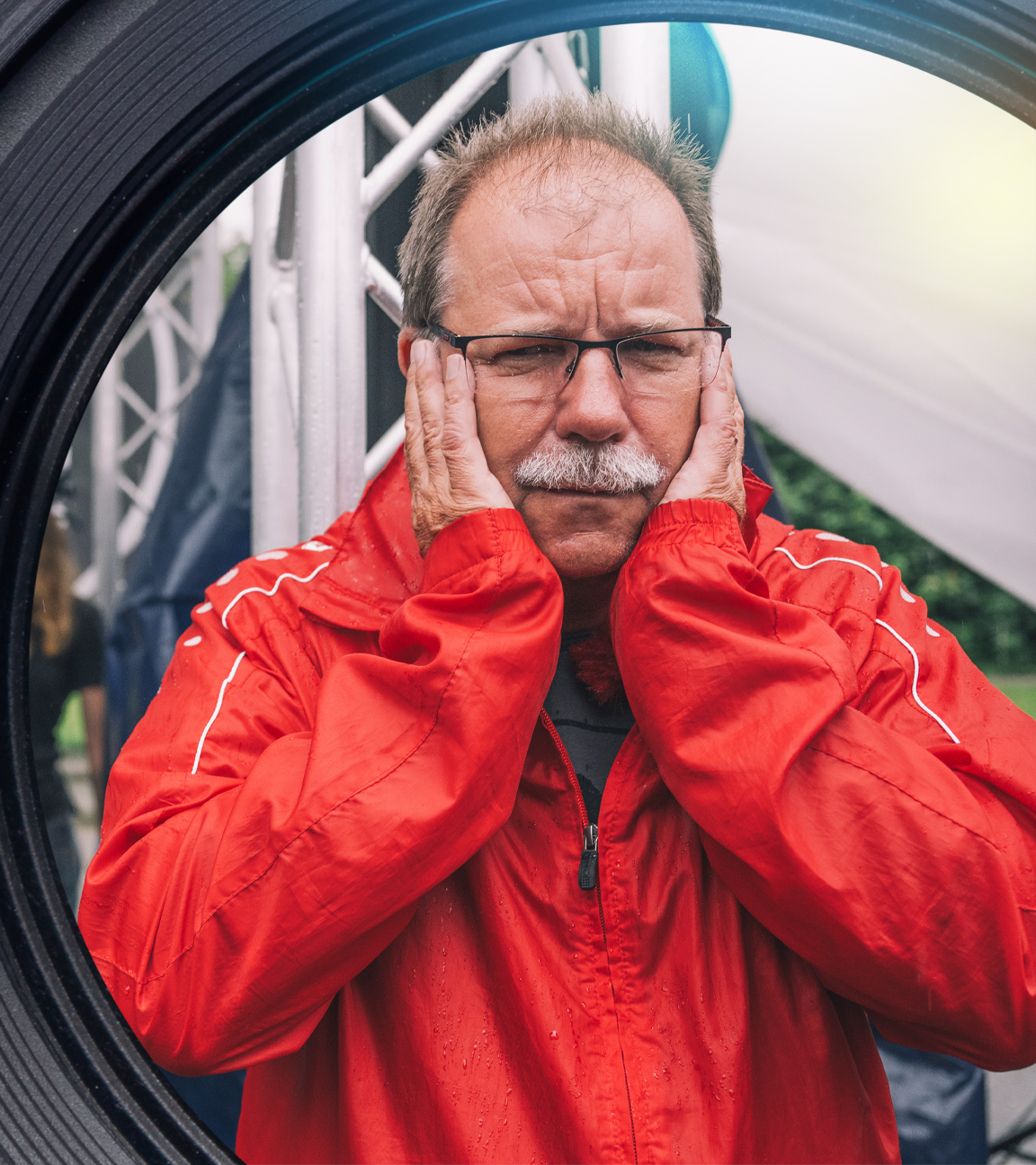 Eine ältere männliche Person mit roter Regenjacke, Brille, Stirnglatze und grauem Schnauzbart steht vor einer großen Lautsprecherbox und hält sich die Ohren zu. Sein Gesichtsausdruck ist gequält. Im Hintergrund stehen drei Personen an einem DJ-Pult.