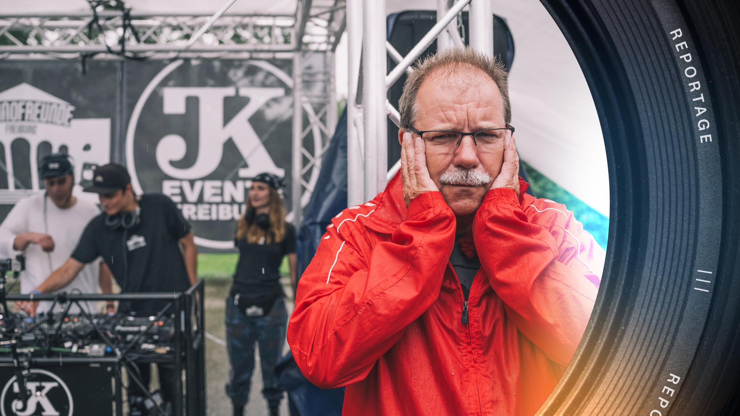 Eine ältere männliche Person mit roter Regenjacke, Brille, Stirnglatze und grauem Schnauzbart steht vor einer großen Lautsprecherbox und hält sich die Ohren zu. Sein Gesichtsausdruck ist gequält. Im Hintergrund stehen drei Personen an einem DJ-Pult.