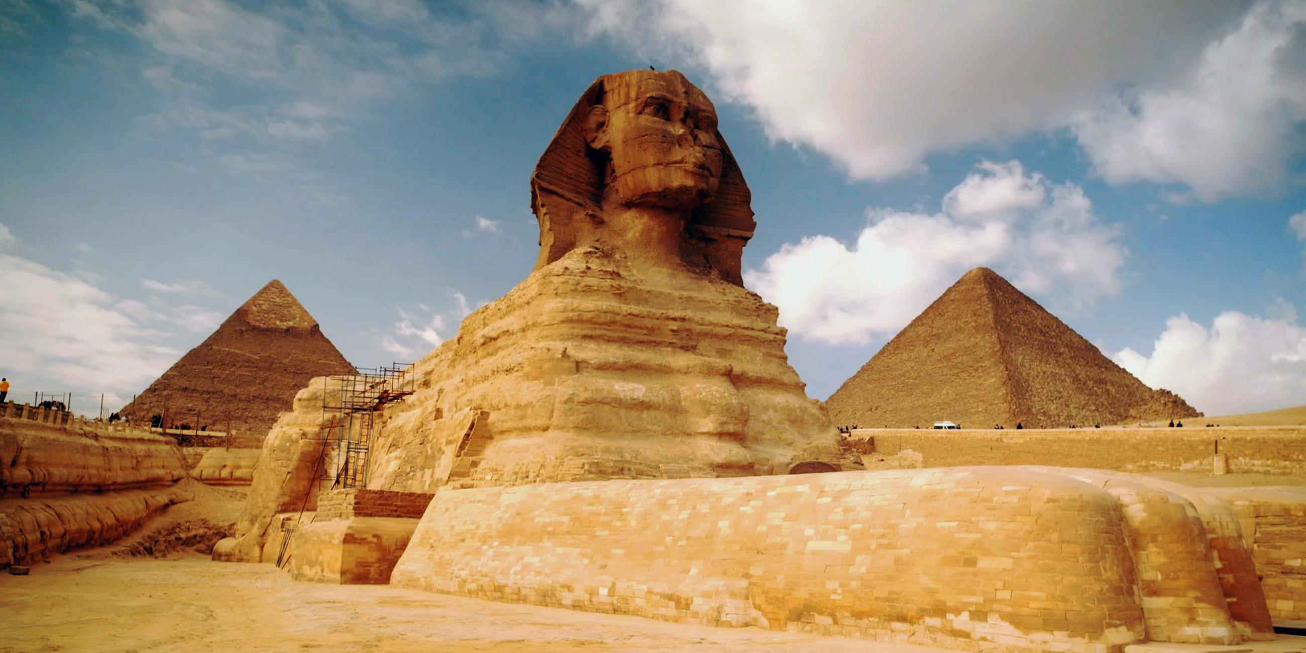 Schrägansicht von unten auf die große Sphinx von Gizeh. An der linken Seite der Sphinx steht ein Baugerüst. Der Himmel ist bewölkt. Im Hintergrund sind zwei Pyramiden zu erkennen.