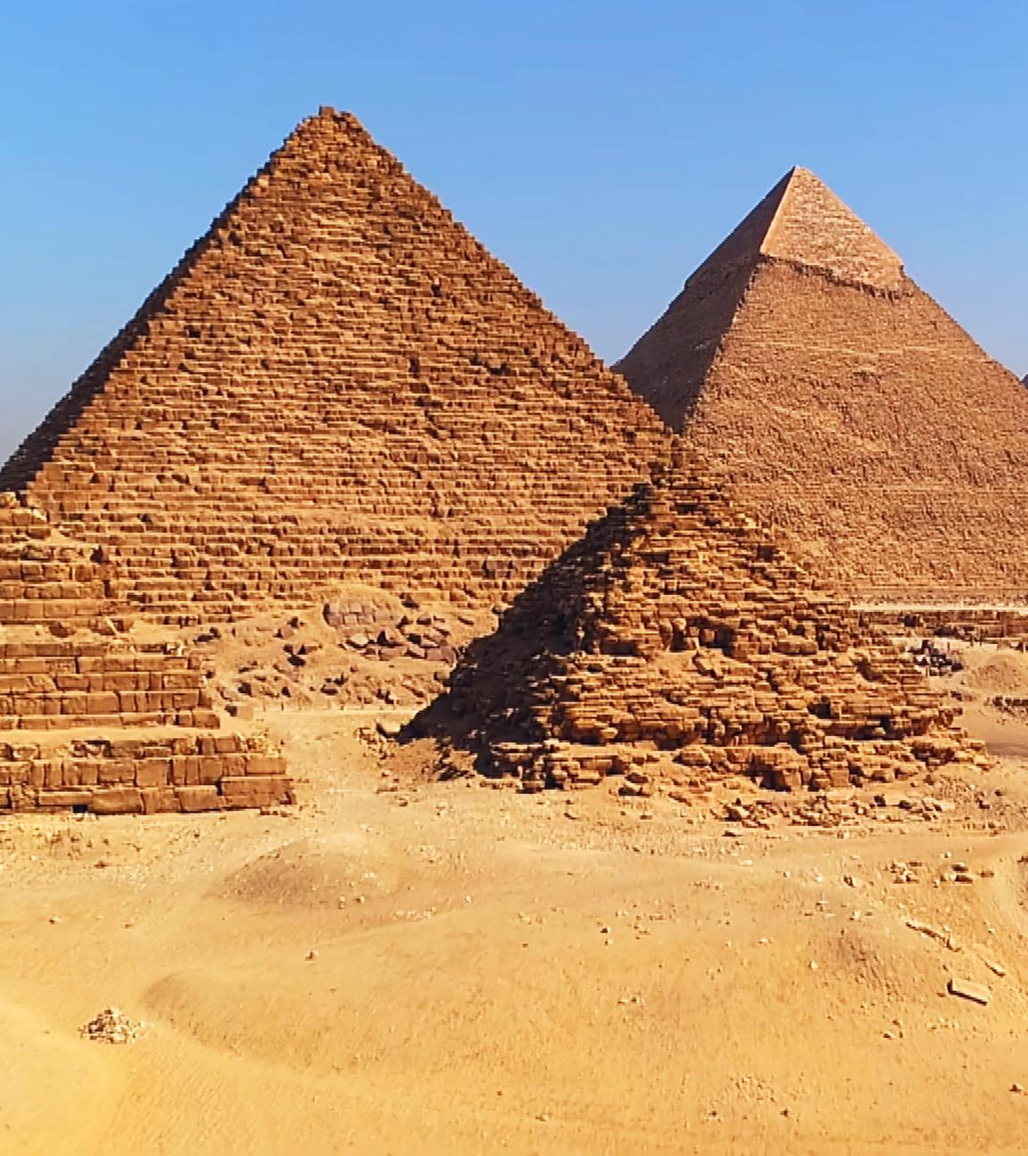 Fünf Pyramiden ragen vor einem trüb blauem Himmel aus einer Sandfläche empor. Im Vordergrund stehen zwei kleine, gleichgroße Pyramiden, eine davon in Stufen. Dahinter, sich überlappend, stehen drei große Pyramiden.