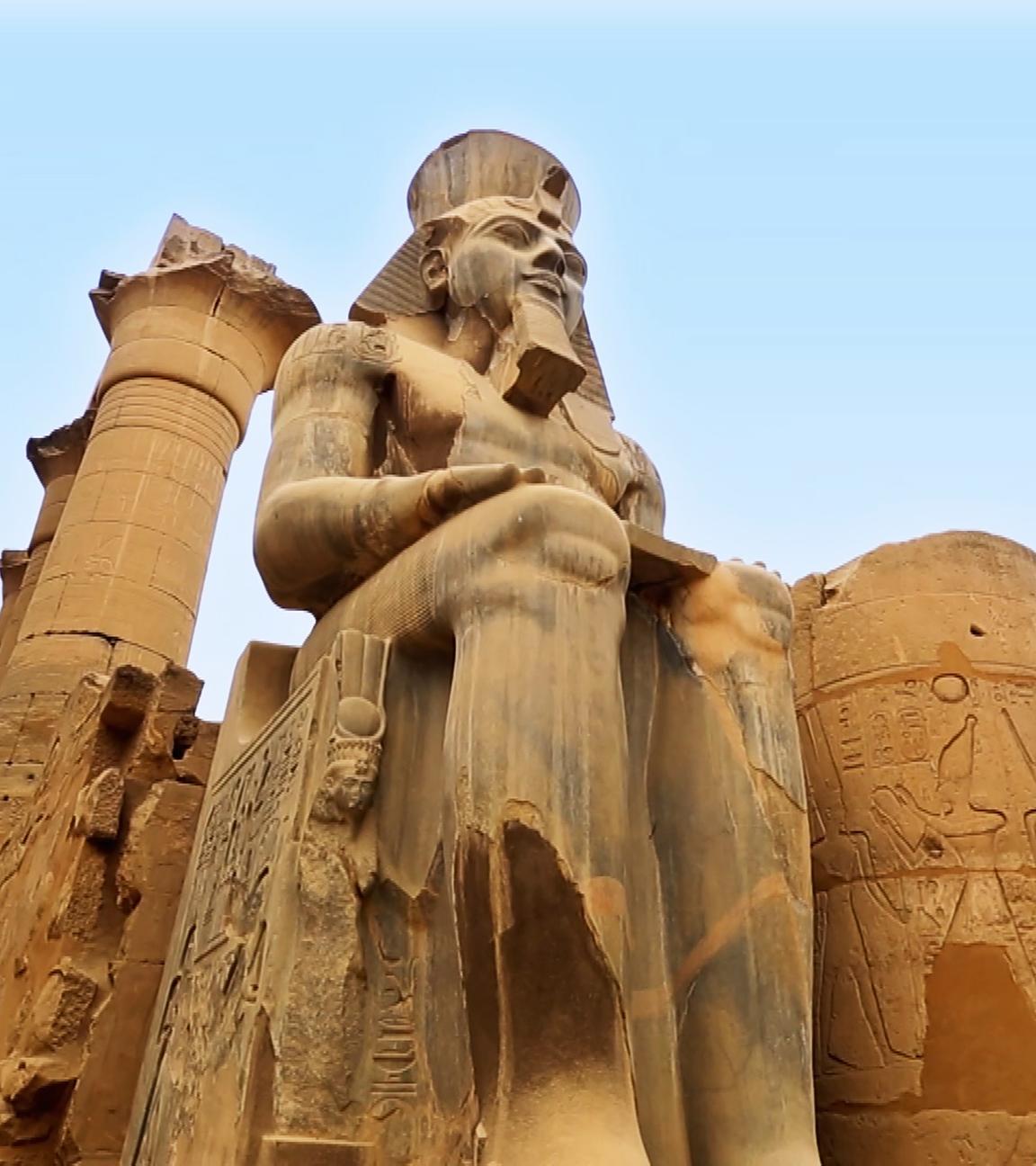 Blick von unten auf eine sitzende Pharaonenstatue: Hände und Unterarme ruhen auf den Oberschenkeln. Auf dem Kopf der Statue ist ein Nemes-Kopftuch mit Uräusschlange, darüber die altägyptische Doppelkrone. Am Kinn trägt die Statue einen Königsbart. Hinter der Statue befindet sich eine Säule mit Hieroglyphen und beschädigtem Kapitell. Im Hintergrund ist blauer Himmel.