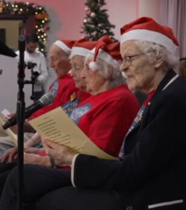 Alte Menschen sitzen mit Weihnachtsmützen auf dem Kopf zusammen und singen Weihnachtslieder.