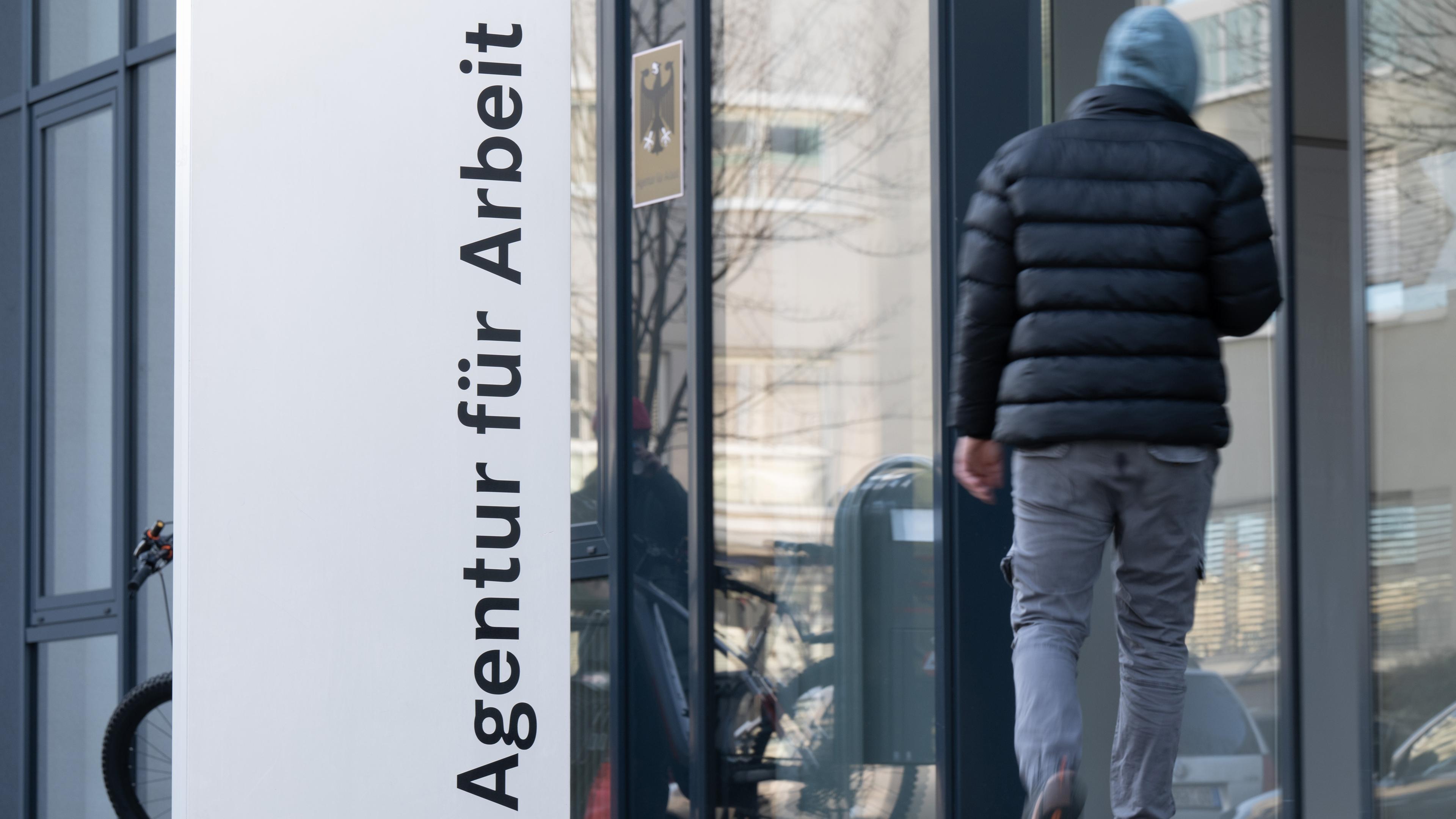 Ein Mann neben einer gläsernen Fassade hinter dem Schriftzug "Agentur für Arbeit".