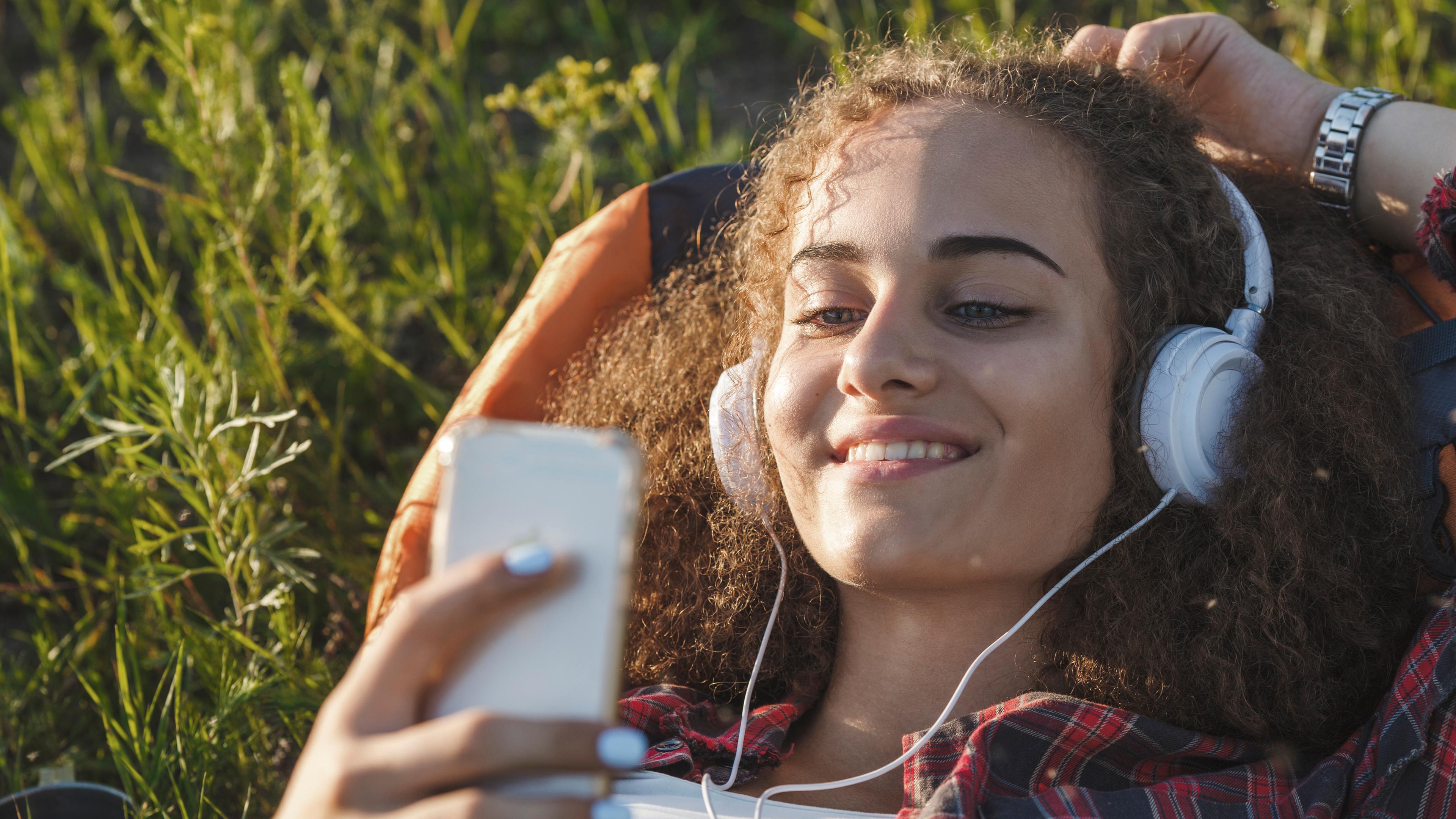 Teenagerin mit Rucksack auf einer Wiese liegend und mit Kopfhörer und Handy Musik hören