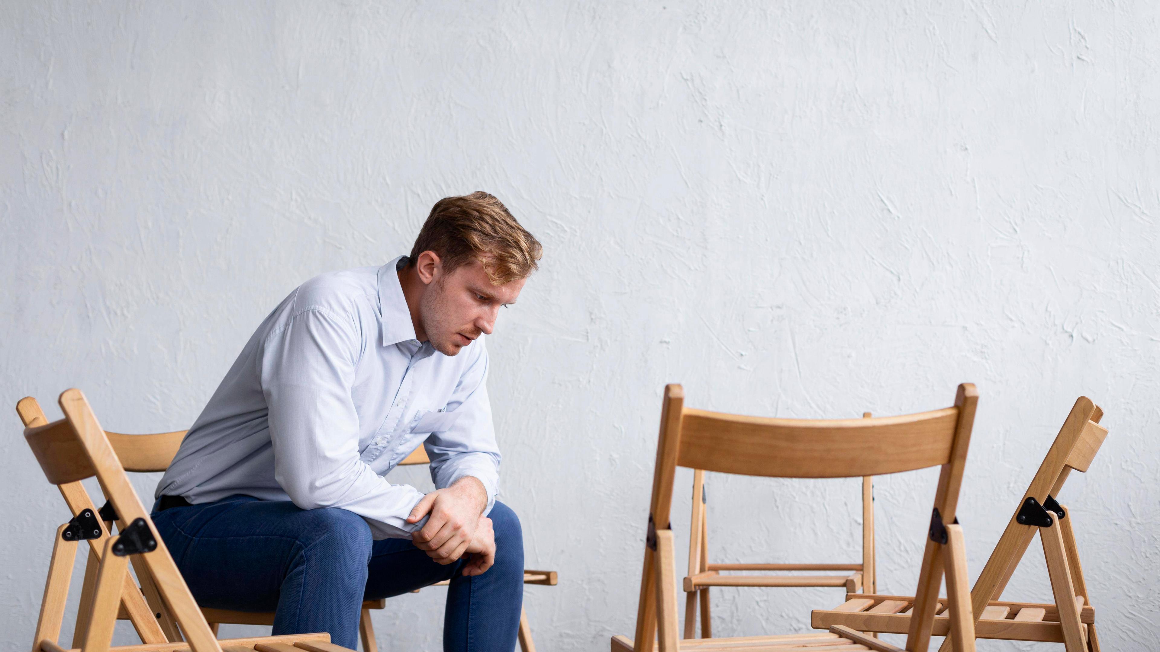 Ein trauriger Mann sitzt in gebeugter Haltung in einer Gruppe aus leeren Stühlen.