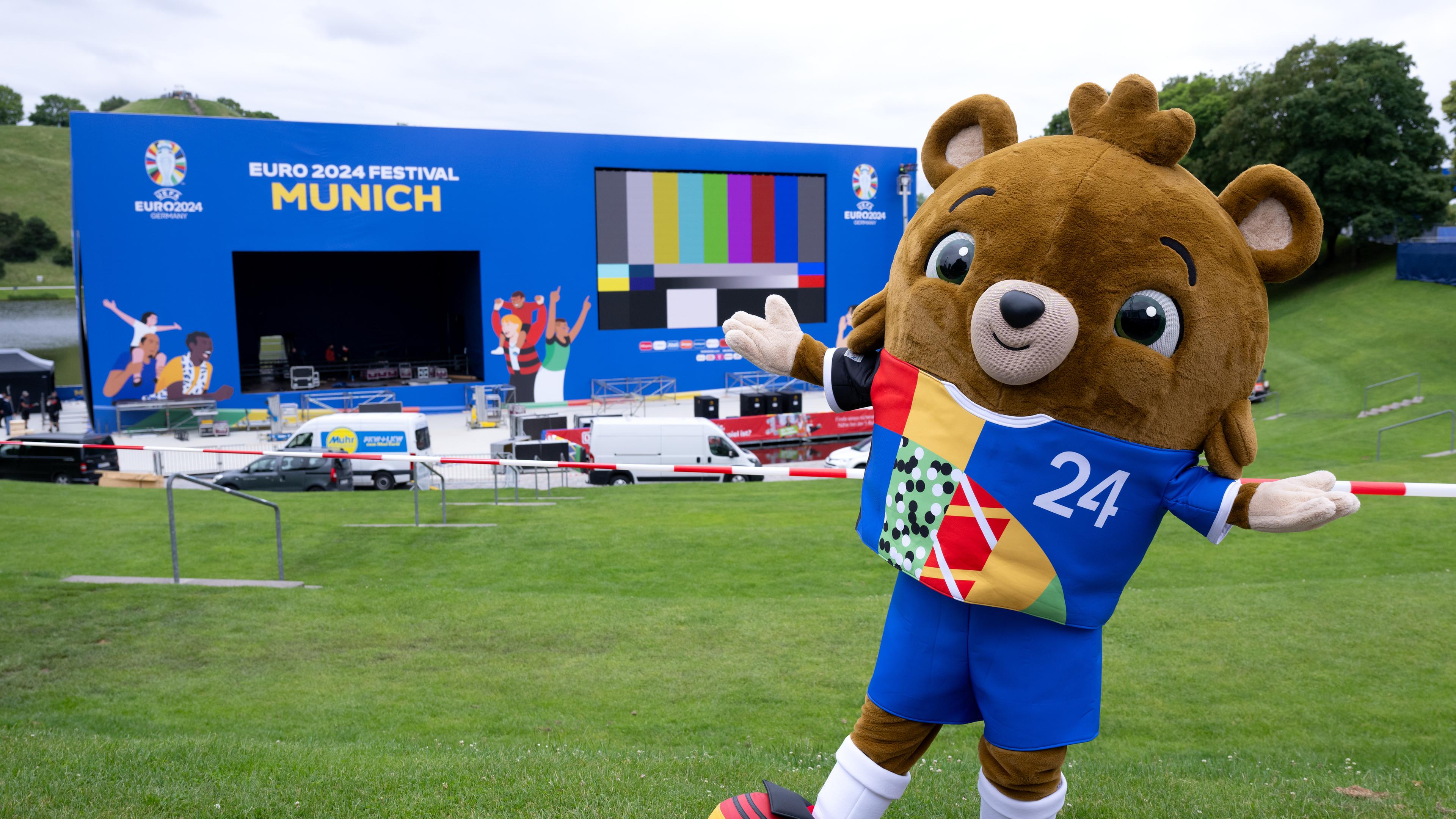 Albärt, das Maskottchen der Fußball-EM 2024, steht im Olympiapark vor einer Leinwand und einer Bühne für die Fan Zone