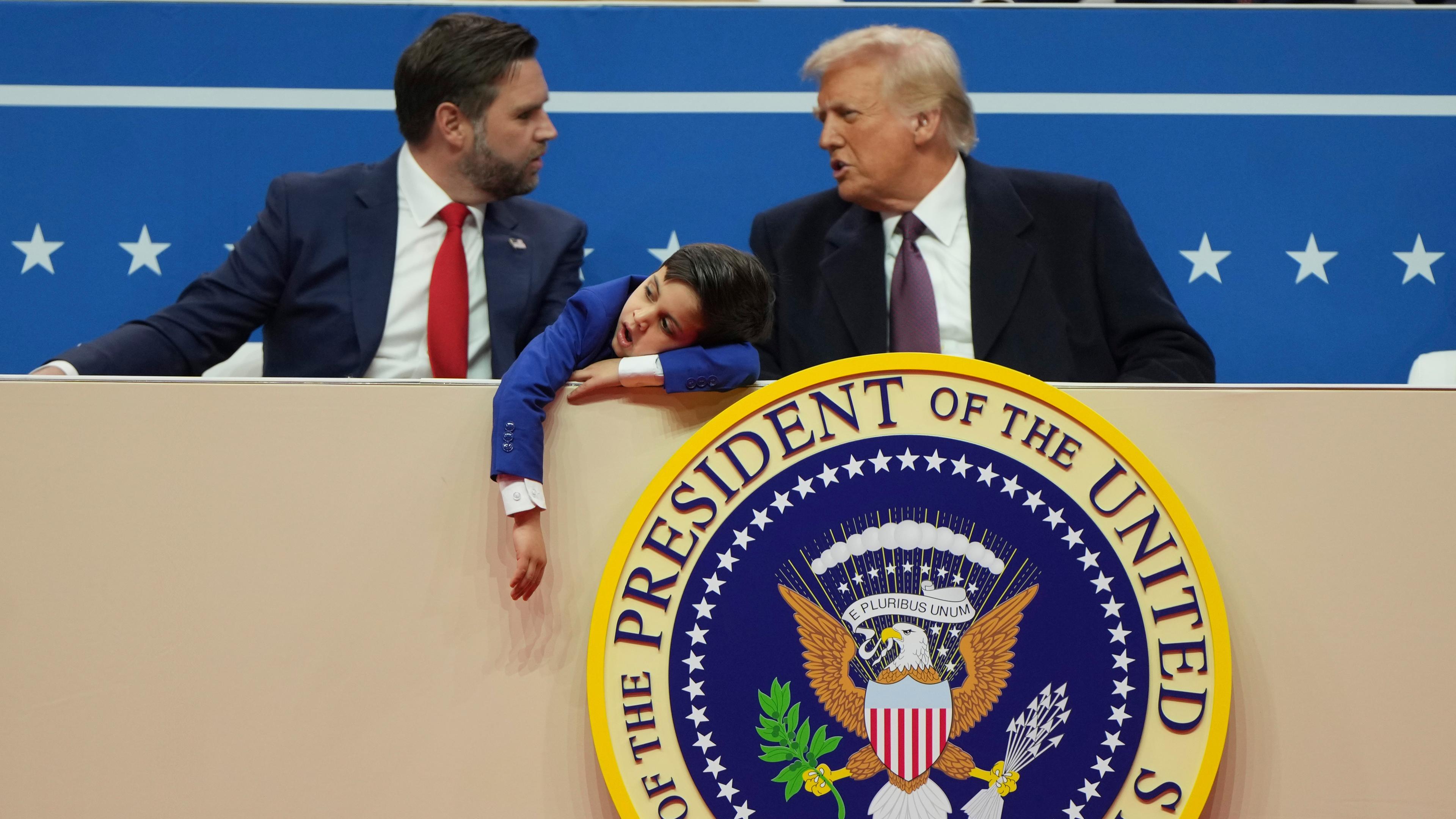 Vizepräsident JD Vance und US-Präsident Donald Trump sitzen auf einer Tribüne vor blauem Hintergrund und unterhalten sich. Zwischen ihnen sitzt der Sohn des Vizepräsidenten in einem blauen Jackett, lässt den Arm über die Tribüne hängen und gähnt. Sein Kopf hat er auf seinen anderen Arm gelegt.