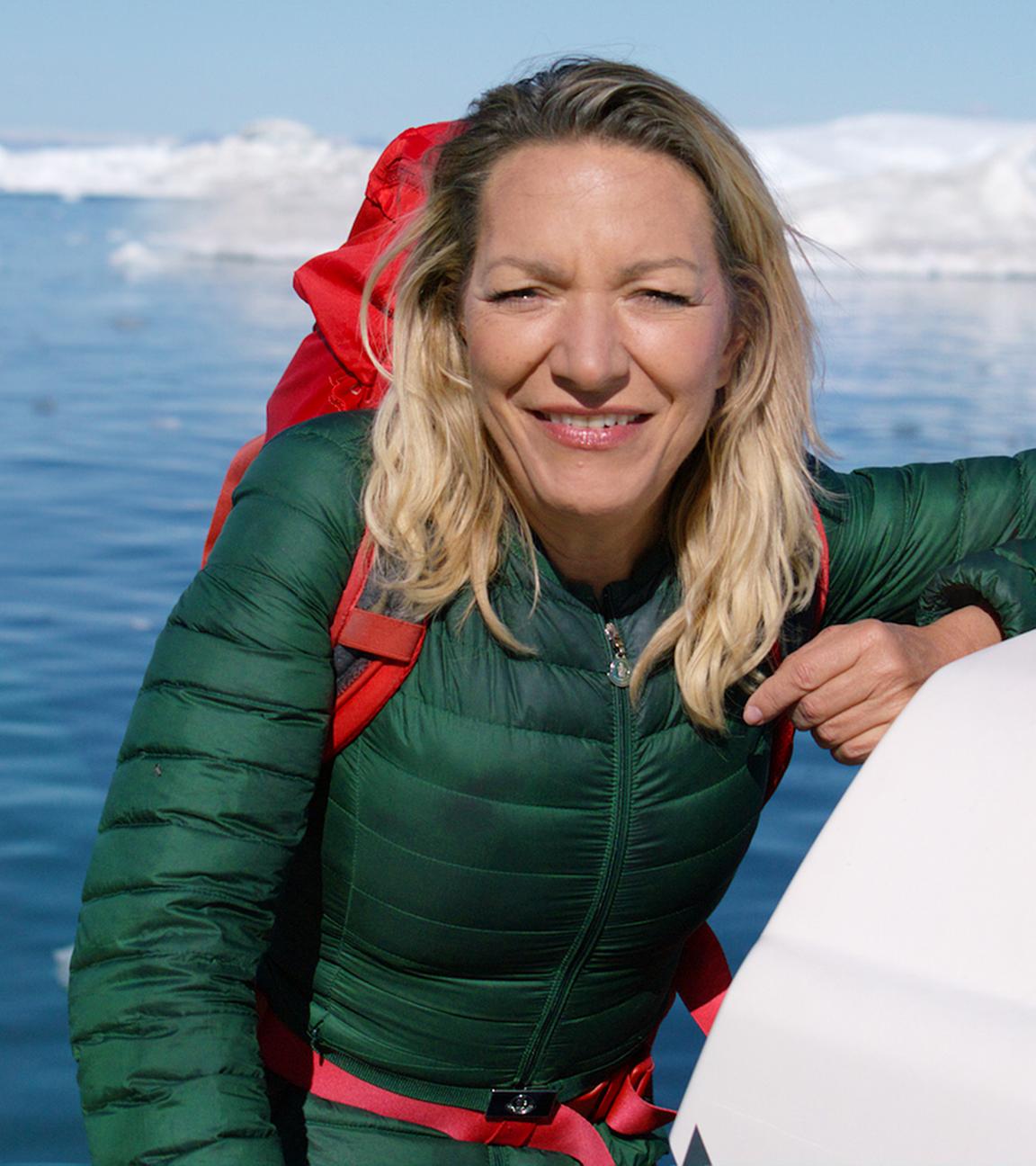 Die Systemforscherin Antje Boetius steht auf einem Boot und schaut lächelnd in die Kamera. Hinter ihr im Wasser fährt ein Schiff zwischen Eisbergen entlang.