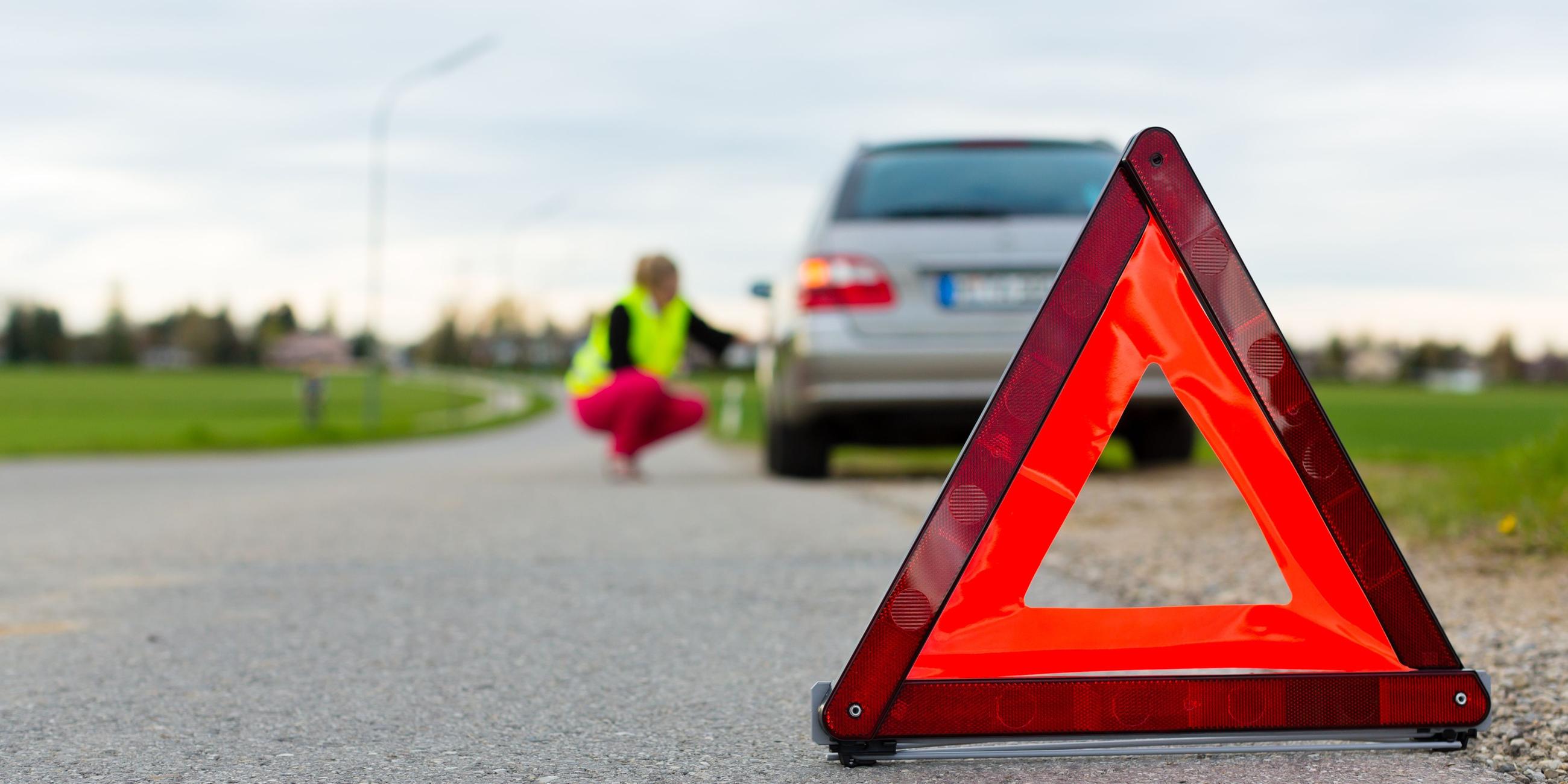 Auf einer Landstraße steht eine Frau mit einer Warnweste neben ihrem Auto, vor dem ein Warndreieck aufgestellt wurde.