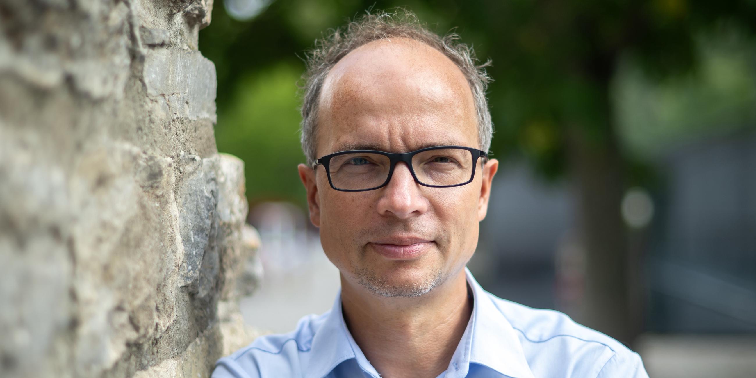 Martin Debes trägt ein hellblaues Hemd und schaut, an eine Steinwand gelehnt, direkt in die Kamera.