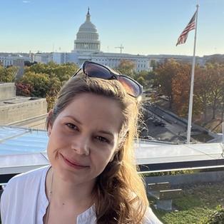 Eine Frau mit braunen Haaren lächelt in die Kamera, im Hintergrund sind Gebäude in Washington DC (USA) zu sehen.