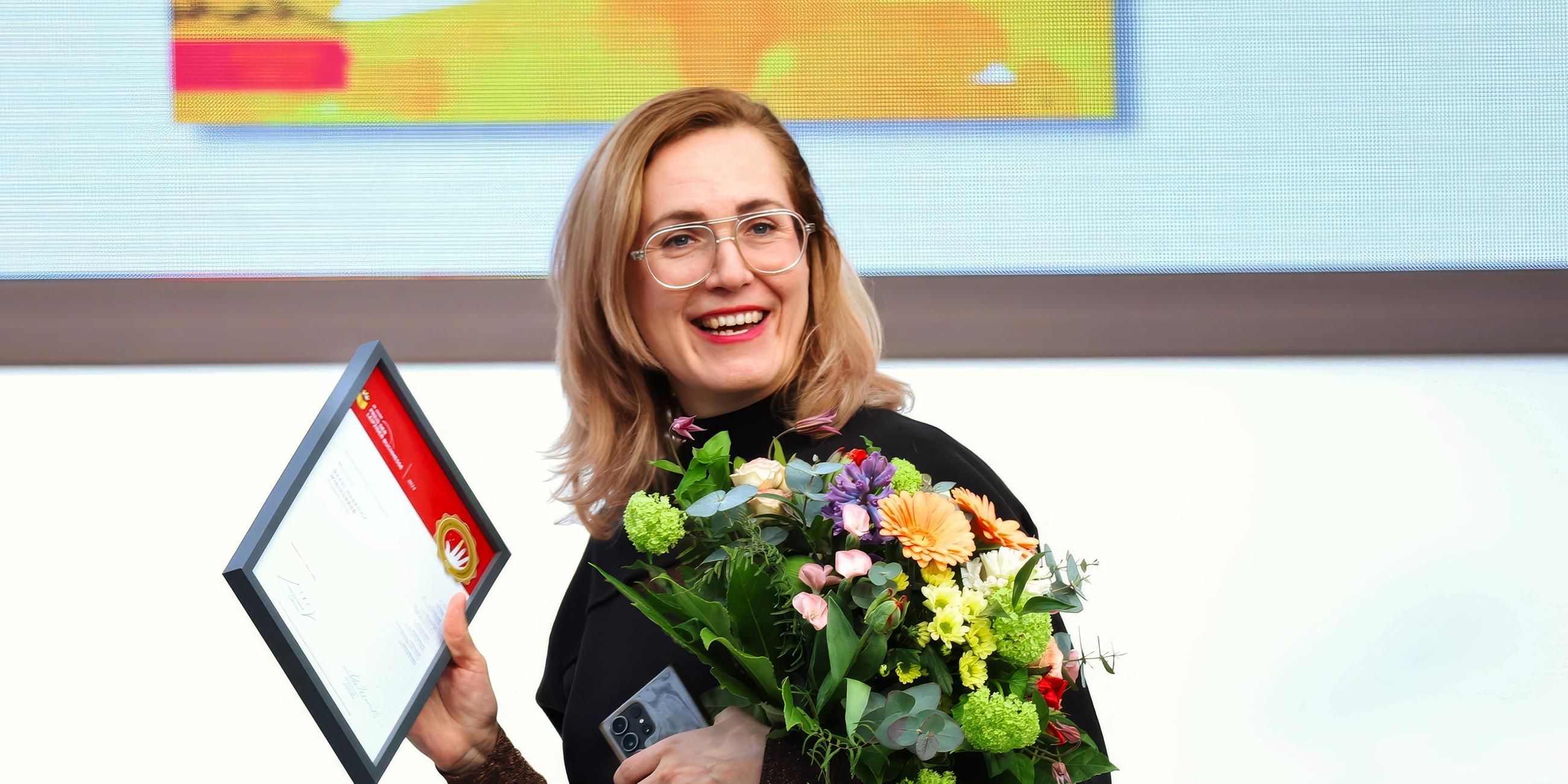 Eine langhaarige blonde Frau hält lachend einen Blumenstrauß und ein gerahmtes Dokument in der Hand.