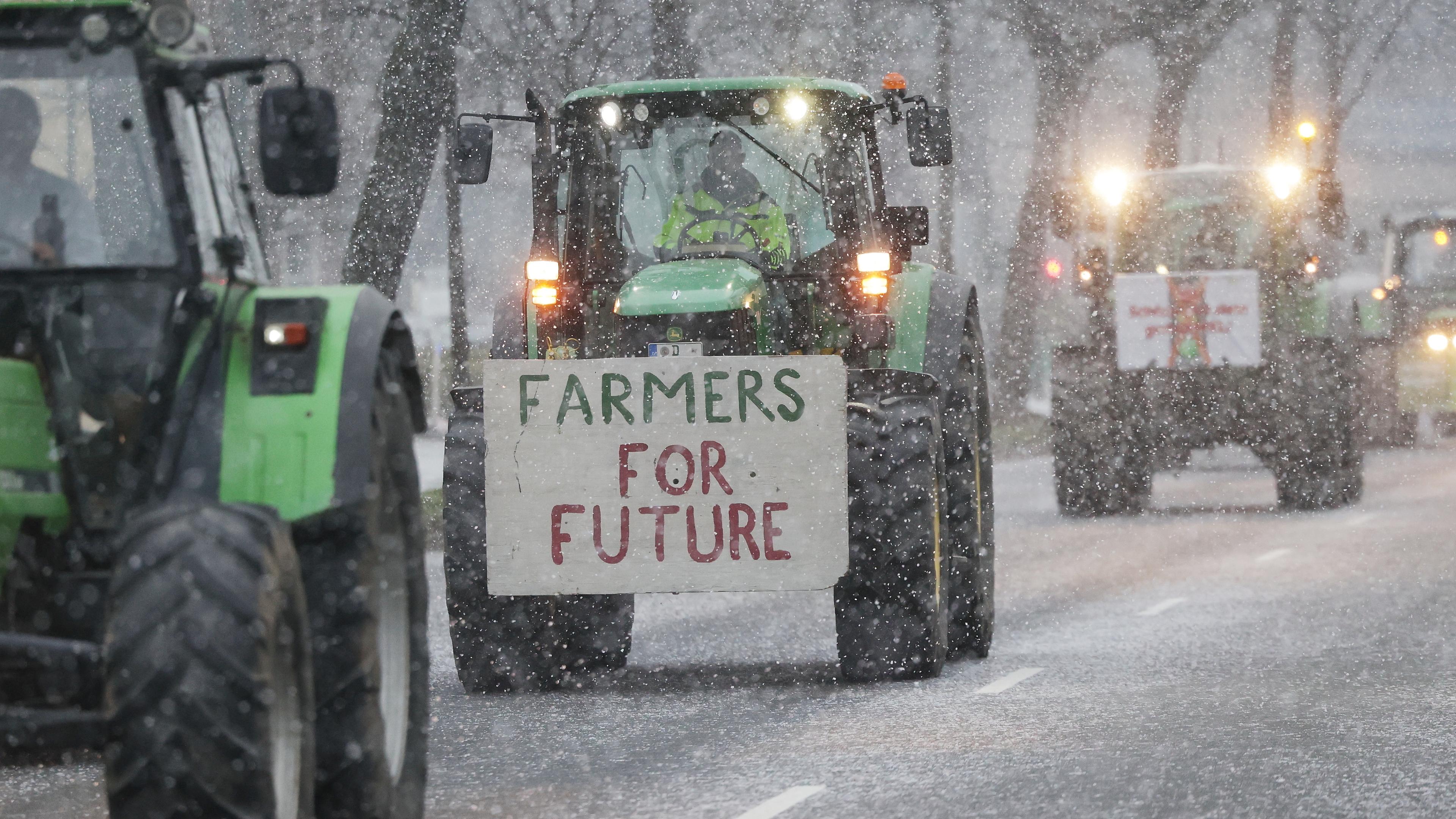 Traktoren fahren bei Schneefall bei einer Demonstration von Bauern über eine Straße. Auf einem Traktor haben Landwirte dabei ein Schild mit der Aufschrift "Farmers For Future" befestigt.