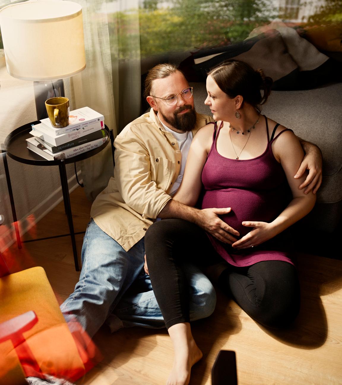 Gitta und Julian sitzen im Wohnzimmer auf dem Boden und schauen sich an. Er hat seine Hand auf ihren Bauch gelegt.