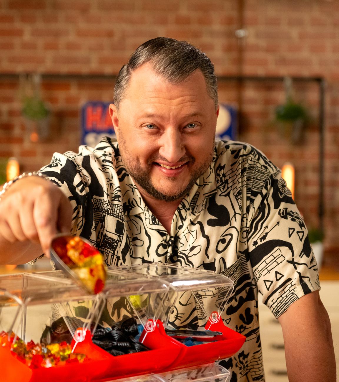 Sebastian Lege steht in seiner Küche hinter einer Süßigkeiten-Theke. In seiner Hand hält er eine Schaufel voll mit Goldbären.