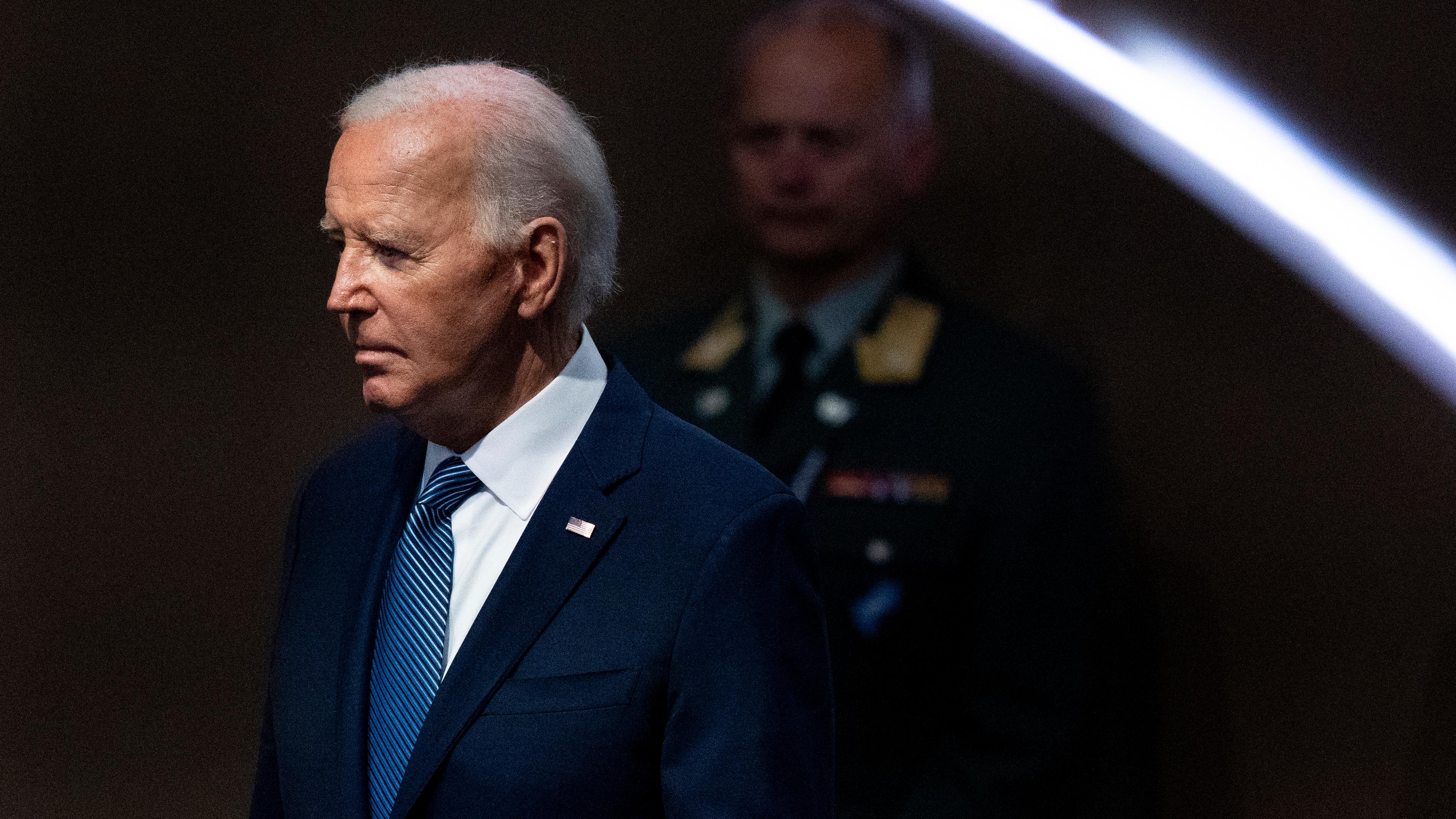 US-Präsident Joe Biden Schaut mit ernster Mine nach links, im Hintergrund ist ein Militär zu sehen. 