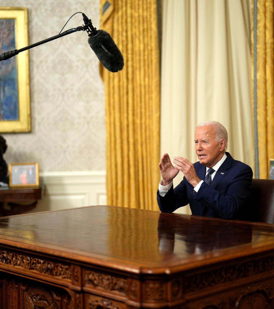 Joe Biden sitzt im Oval Office hinter seinem Schreibtisch und spricht fokussiert in Richtung der Kameras. Ein Mikrophonständer ist neben dem Schreibtisch zu sehen.