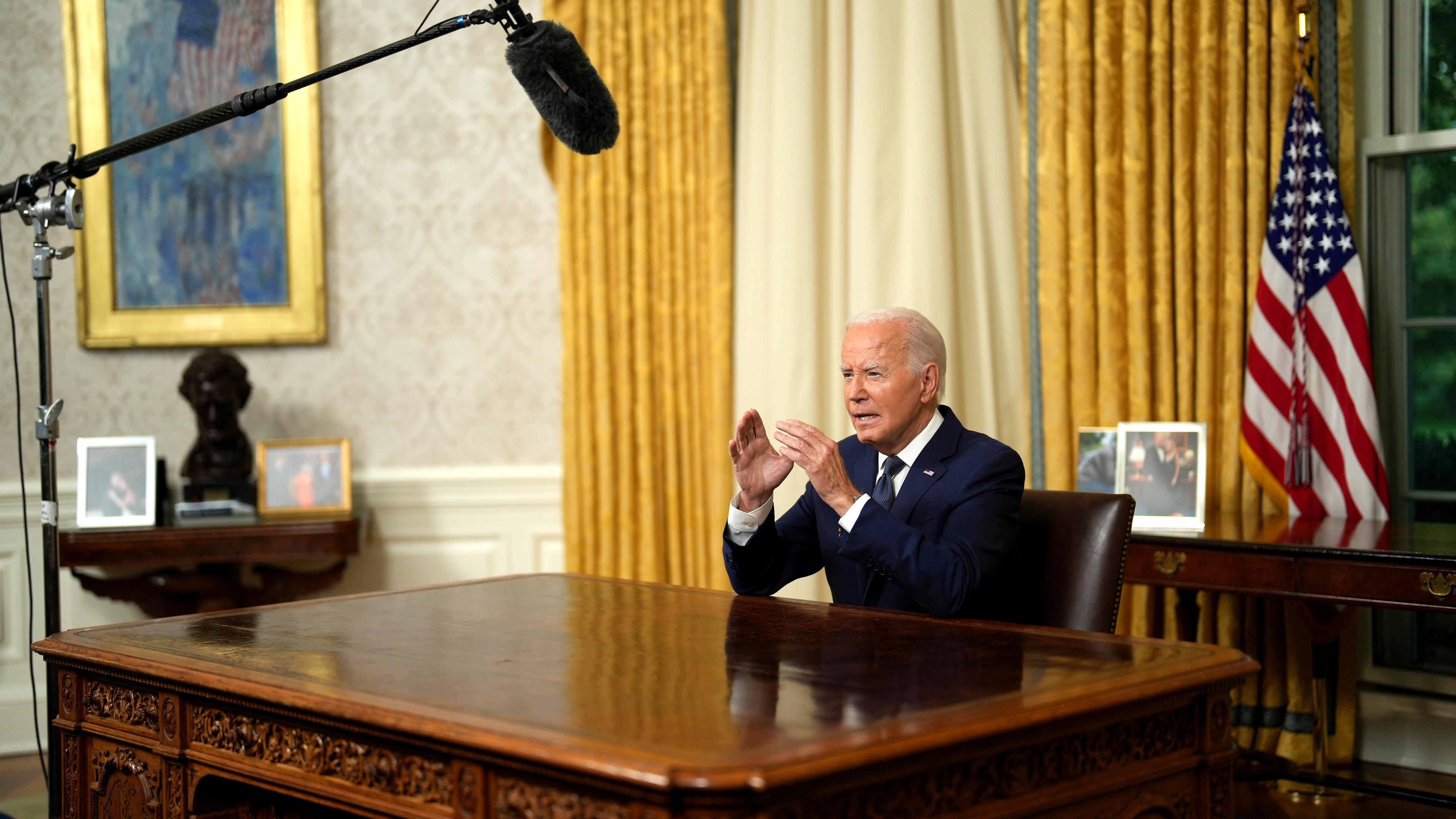 Joe Biden sitzt im Oval Office hinter seinem Schreibtisch und spricht fokussiert in Richtung der Kameras. Ein Mikrophonständer ist neben dem Schreibtisch zu sehen.