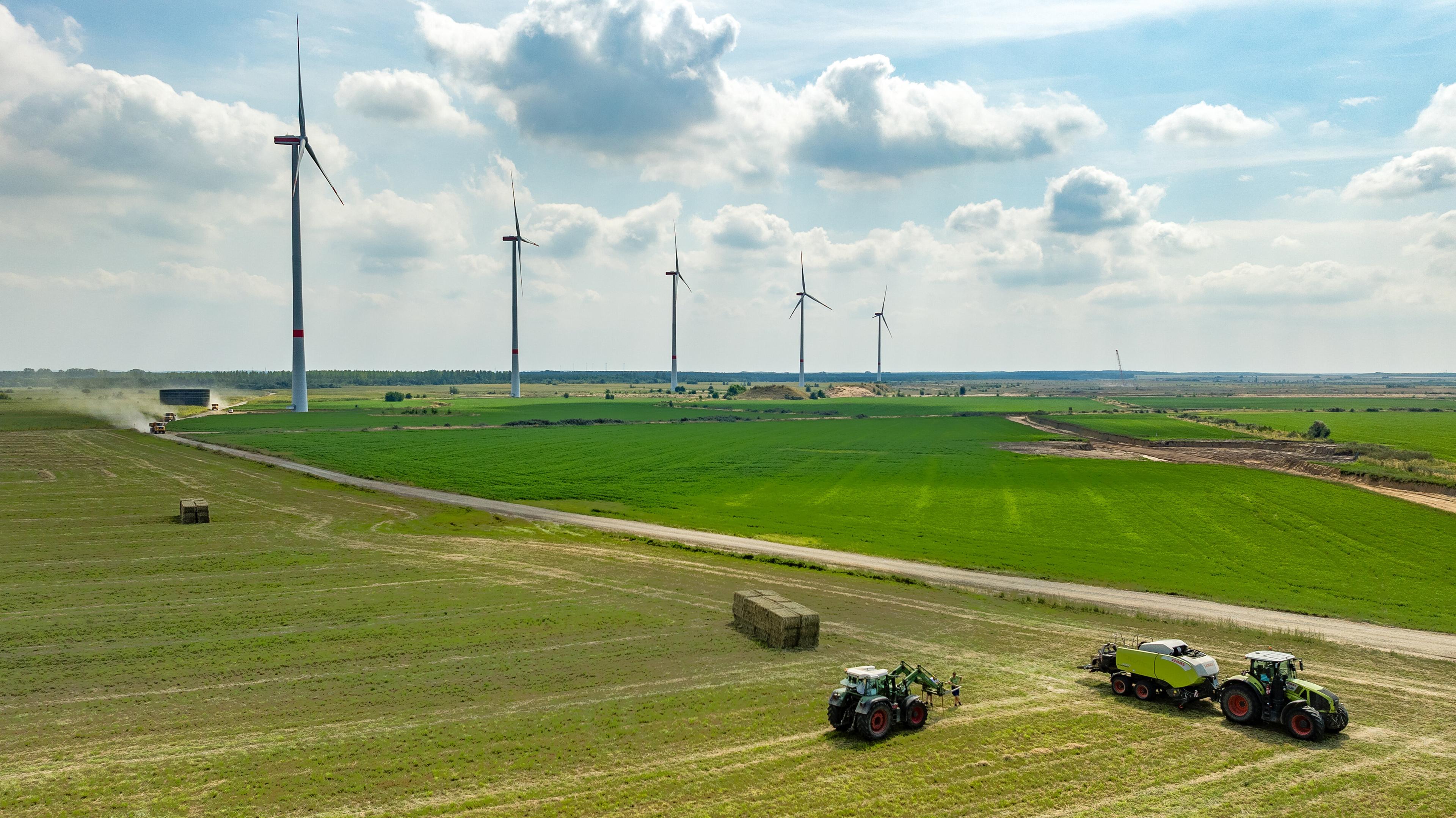 Hinter landwirtschaftlichen Maschinen, mit denen ein Feld bearbeitet wird, stehen eine Reihe von Windrädern.