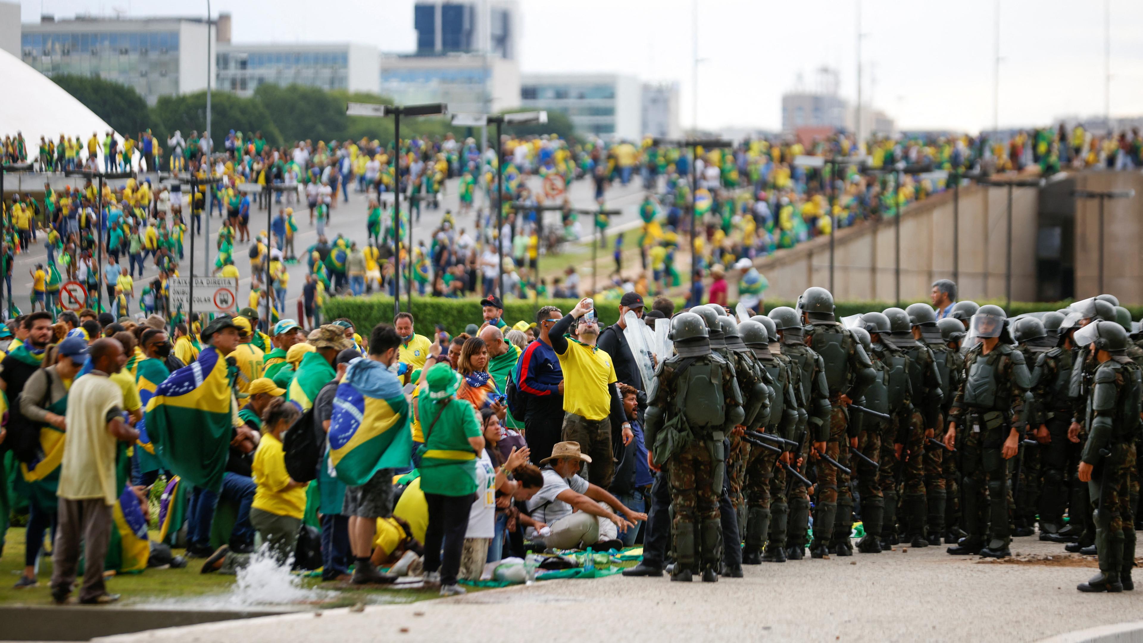 Polizisten in Uniform und Helmen stehen Protestierenden mit Brasilien Flaggen gegenüber.