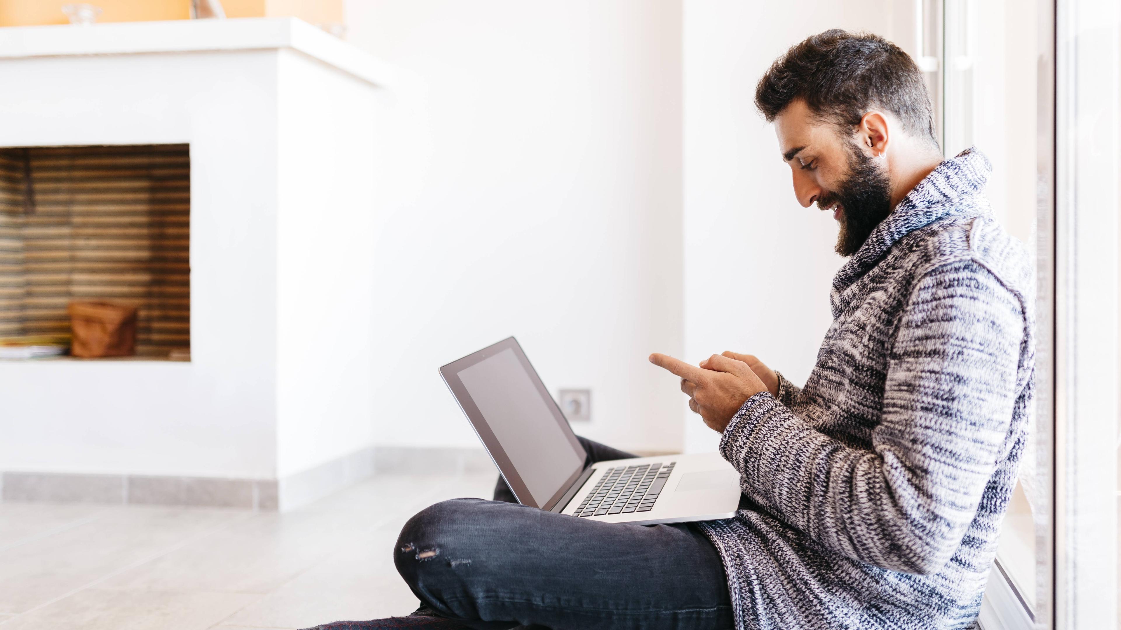 Ein Mann sitzt mit einem Laptop und einem Smartphone auf dem Boden vor einem Kamin. 