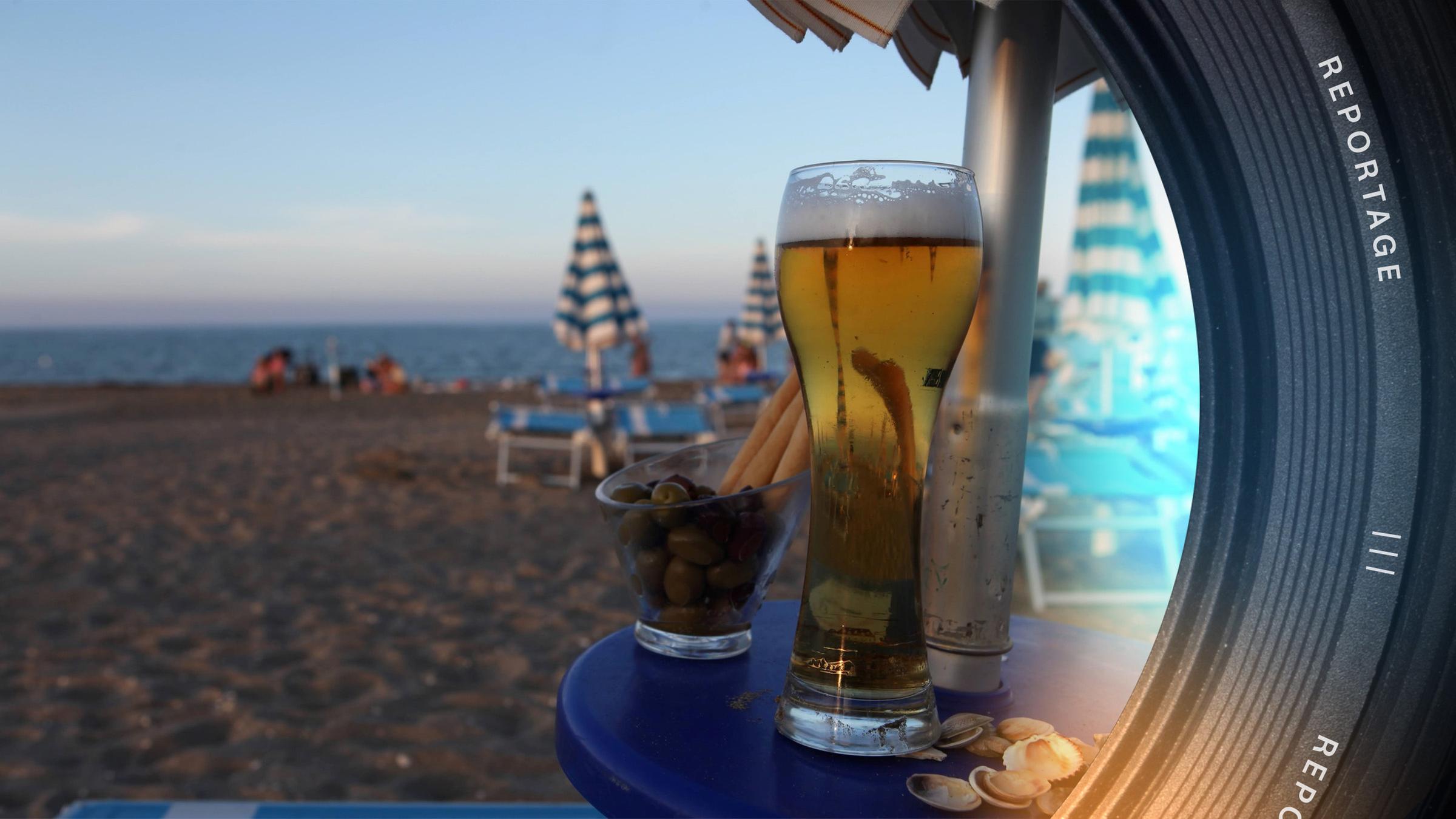 Auf einem blauen Tisch steht im Vordergrund rechts ein volles Weizenbierglas. Im Hintergrund befinden sich der Strand in der dämmrigen Abendsonne, Strandliegen und zugeklappte Sonnenschirme.