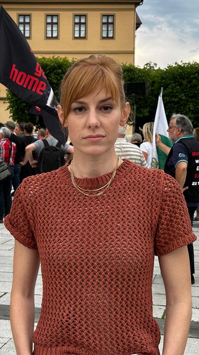 "Sachsen, warum bist du so gespalten?": Eva Schulz bei einer Demo in Sachsen: Sie steht auf einem Platz und schaut in die Kamera. Hinter ihr stehen viele Menschen, eine Person hält eine Deutschlandfahne, auf einer anderen steht "Ami go home".