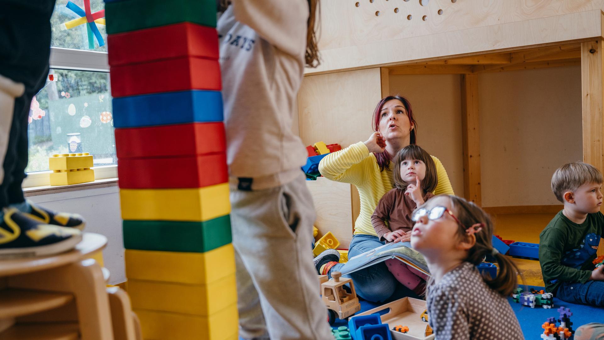 "Kein Kinderspiel! - System Kita am Anschlag": Im Kindergarten: Erzieherin Diana sitzt mit ihren Kindergartenkindern am Boden, zwei Kinder stehen im Vordergrund und bauen einen Turm.