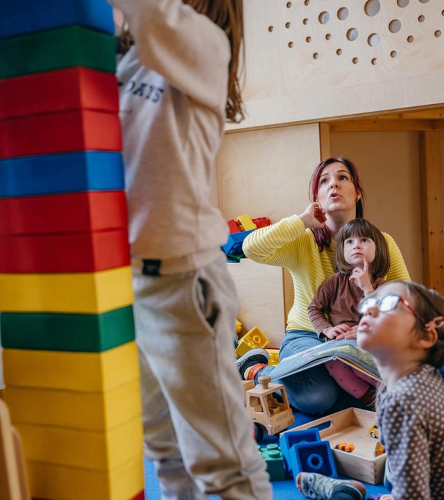 "Kein Kinderspiel! - System Kita am Anschlag": Im Kindergarten: Erzieherin Diana sitzt mit ihren Kindergartenkindern am Boden, zwei Kinder stehen im Vordergrund und bauen einen Turm.