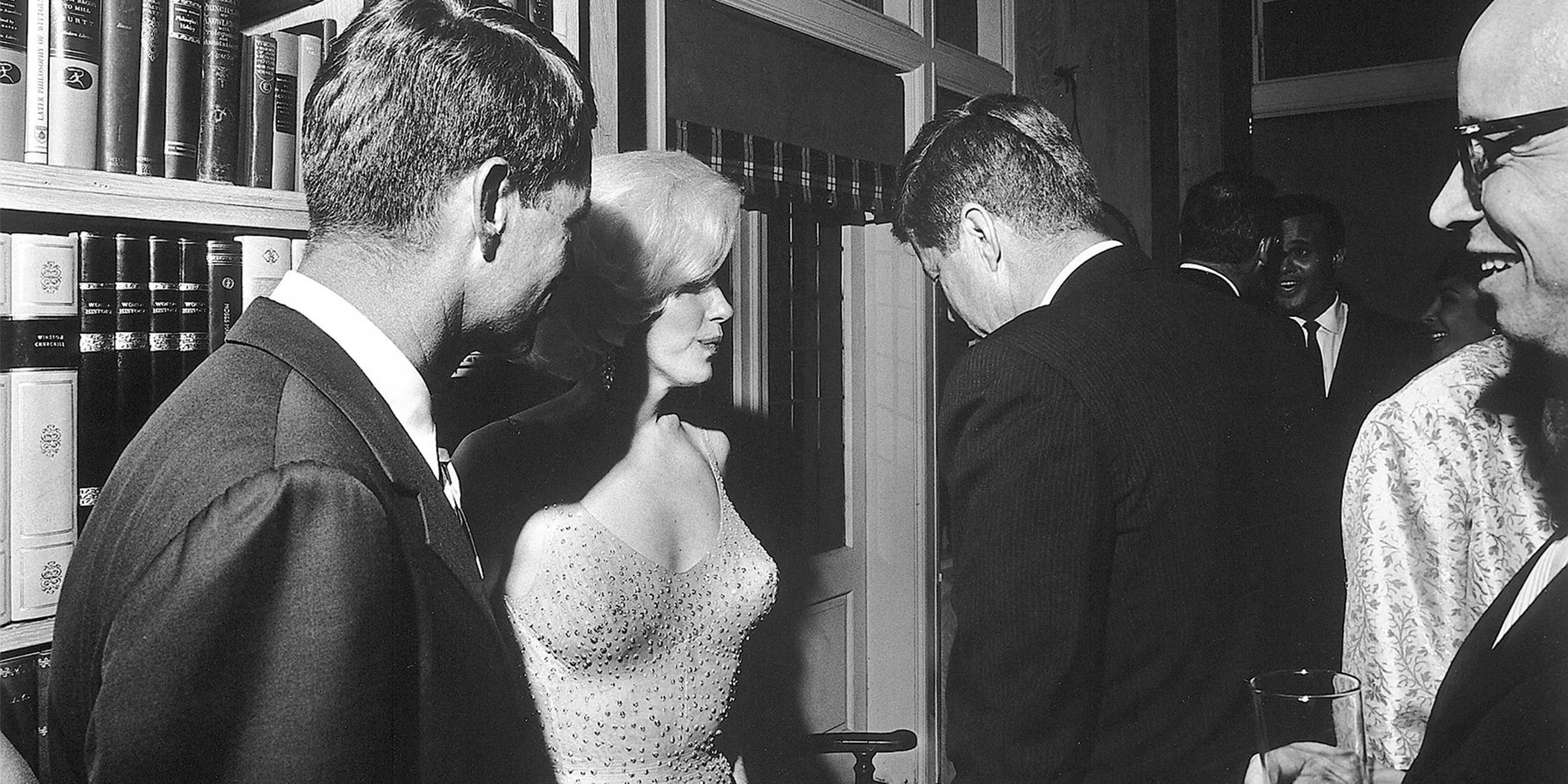 Schwarz-Weiß-Foto: Marilyn Monroe steht zusammen mit John F. Kennedy und Robert Kennedy auf einer Party.