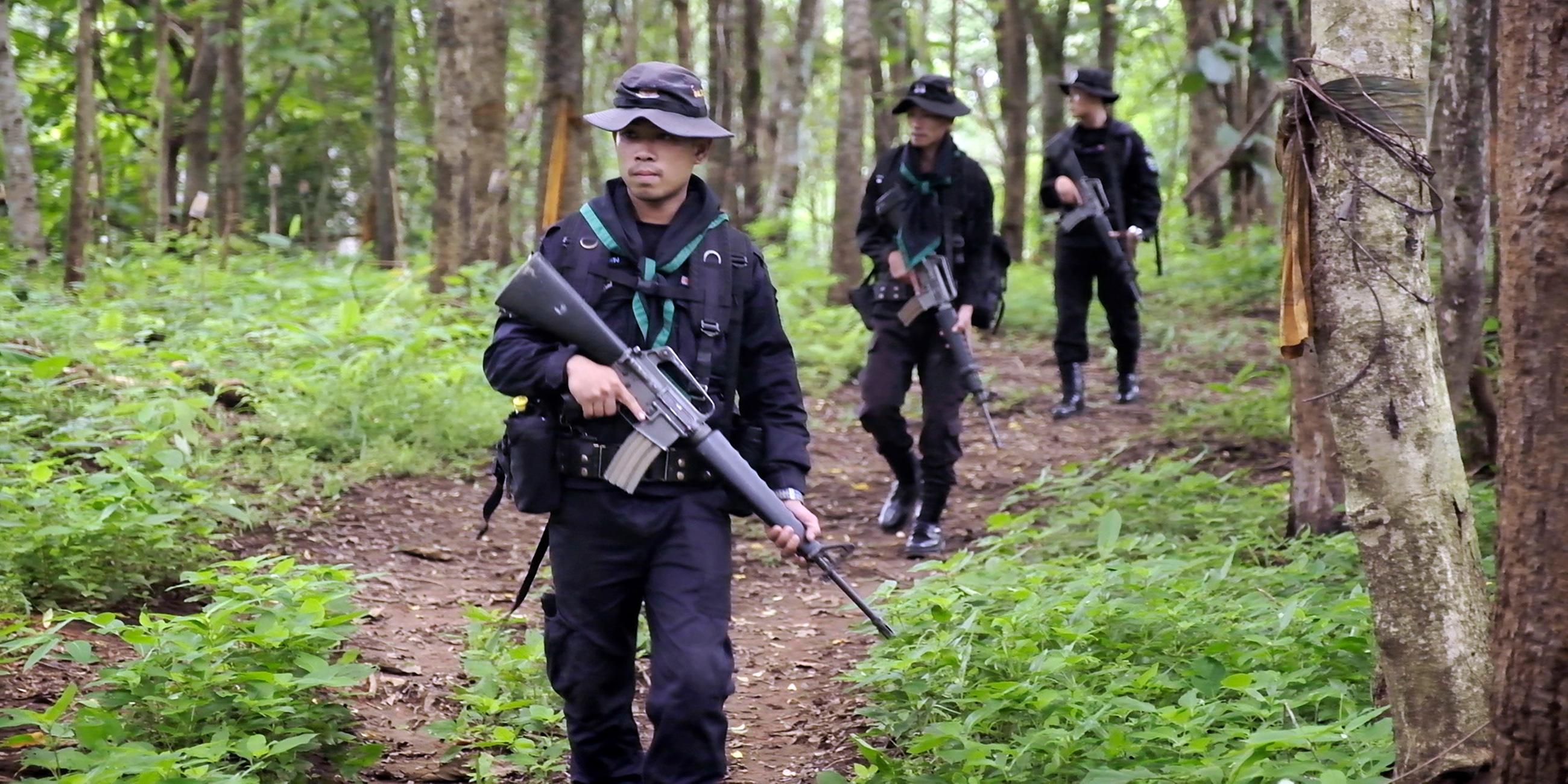 Drei Männer mit Hüten und schwarzen Uniformen laufen hintereinander über einen Waldweg. Alle drei halten schwere Maschinengewehre in den Händen.