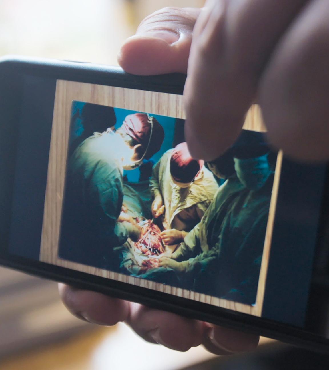 Nahaufnahme eines Smartphones. Auf dem Gerät ist ein Foto zu sehen, das mehrere Ärzte in grünen Kitteln und roten Hauben bei einer Operation zeigt.