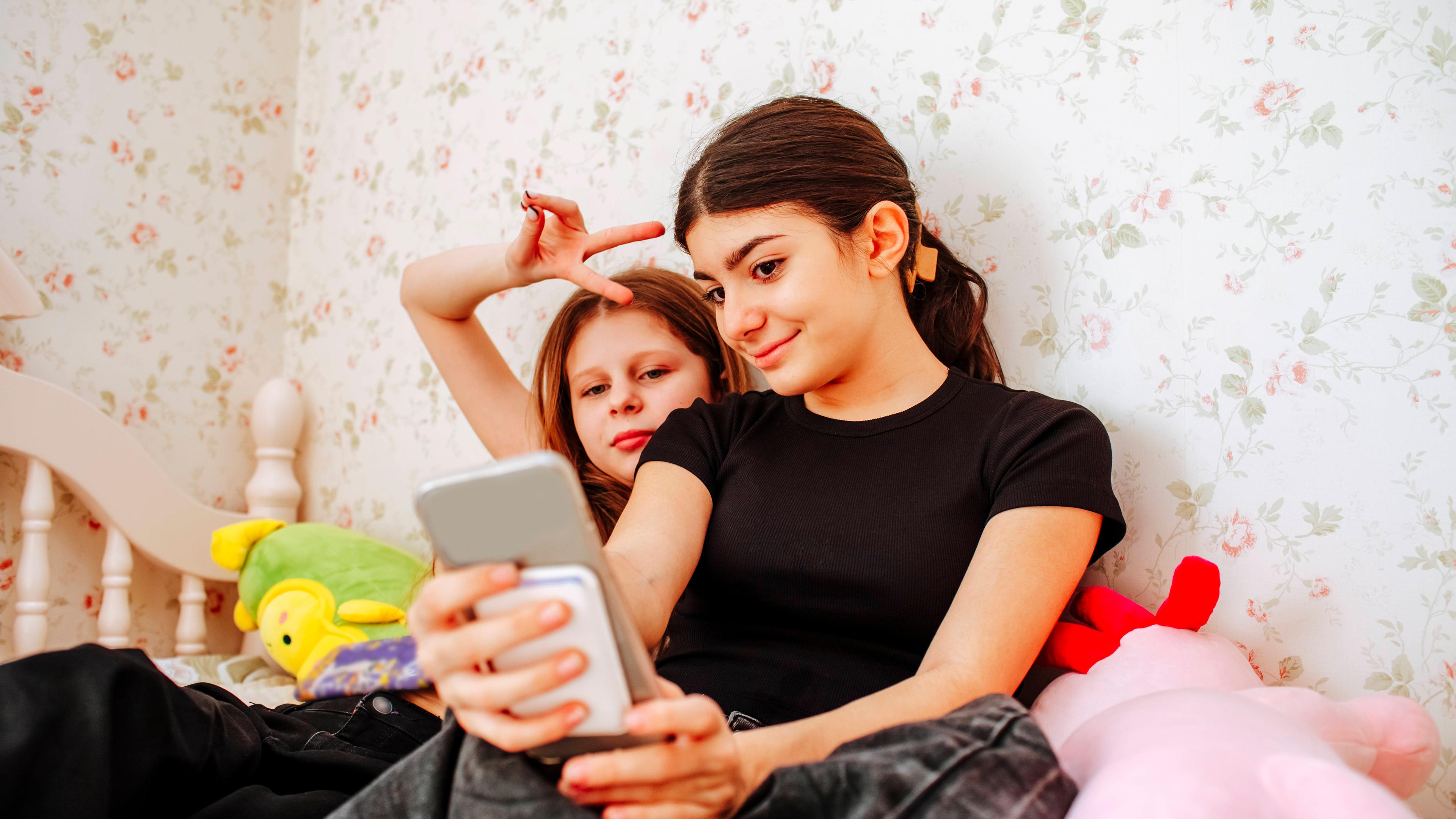 Zwei Mädchen sitzen auf dem Bett im Kinderzimmer und nehmen ein Selfie auf.