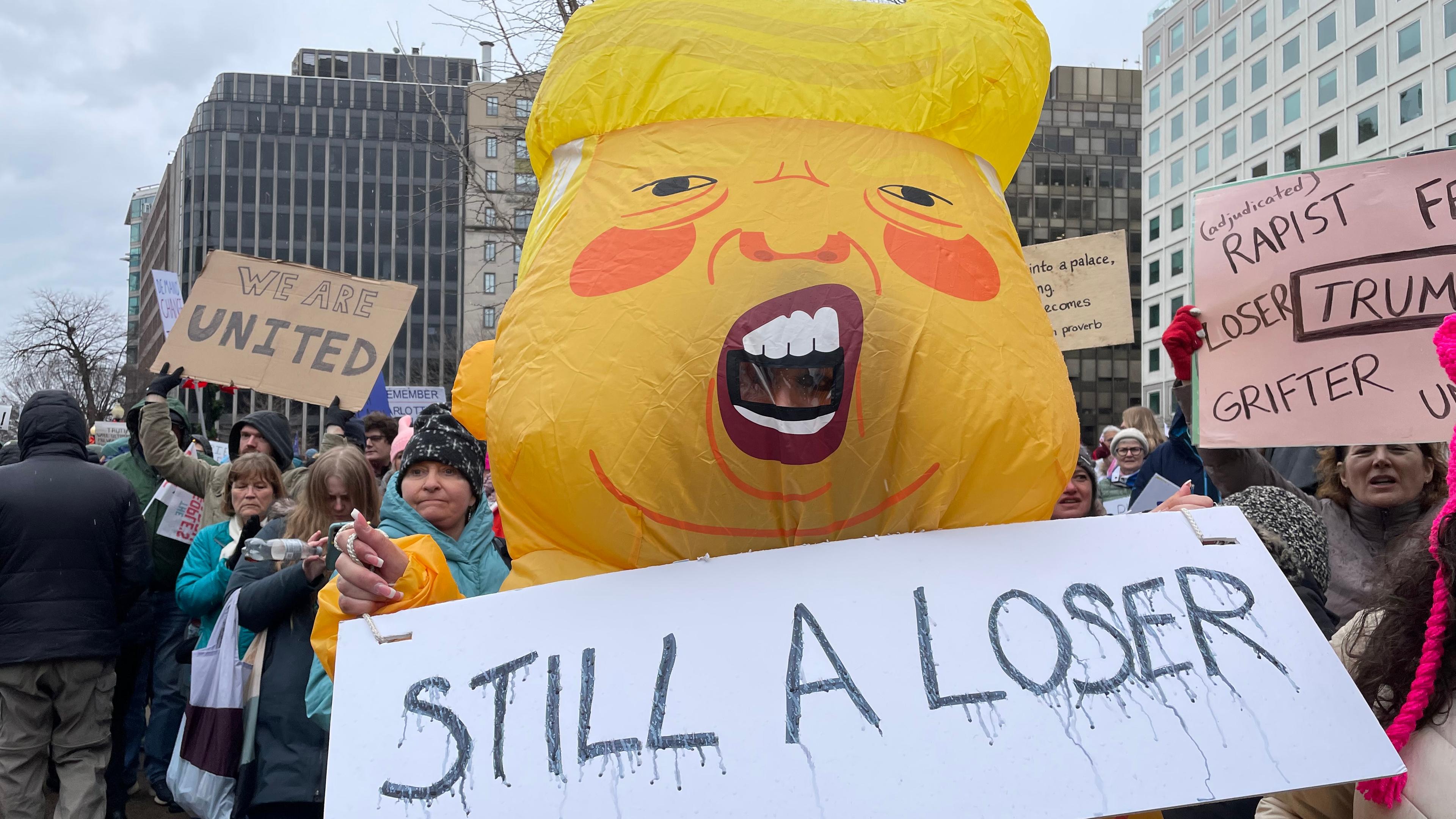 Eine Frau steht in einem aufgeblasenen Trump-Ballon-Kostüm auf einer Demo. Sie hält ein Schild in der Hand mit der Aufschrift: Noch immer ein Verlierer.