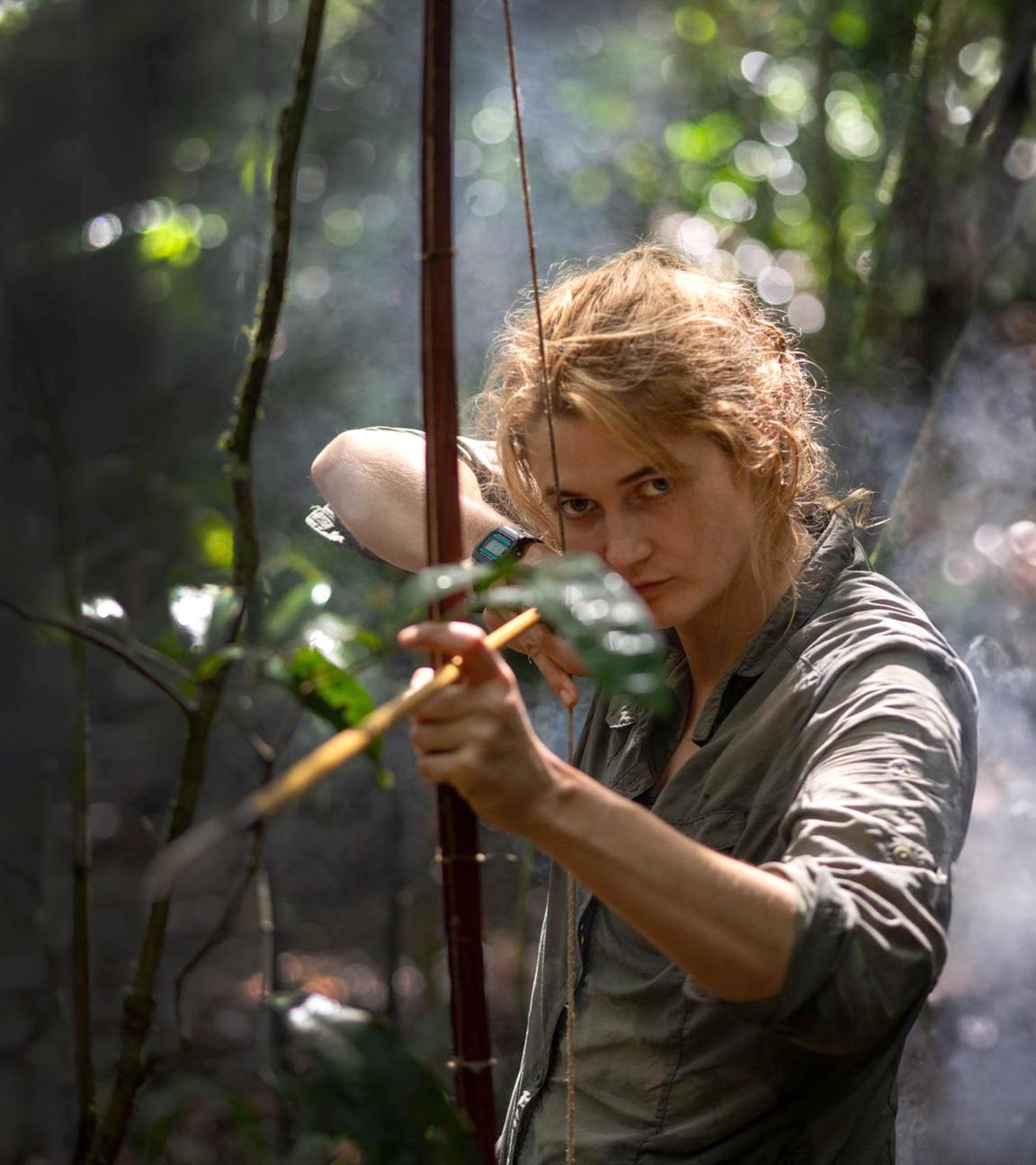 Lucy Shepherd steht im dichten Urwald und hält Pfeil und Bogen in der Hand. Mit der rechten Hand hat sie einen Pfeil aufgezogen und fokussiert ein Ziel.