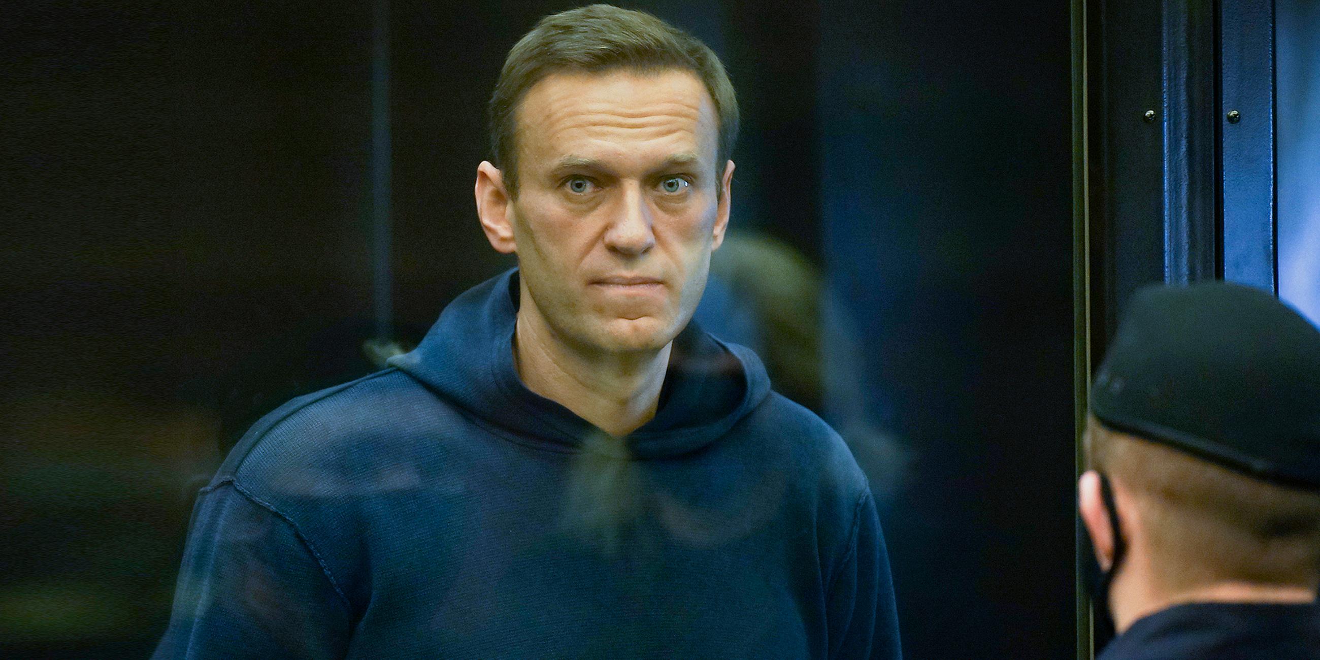 Alexey Nawalny steht im Käfig im Gericht, bekleidet mit einem dunkelblauen Hoody und schaut direkt durch die Glasscheibe in die Kamera.