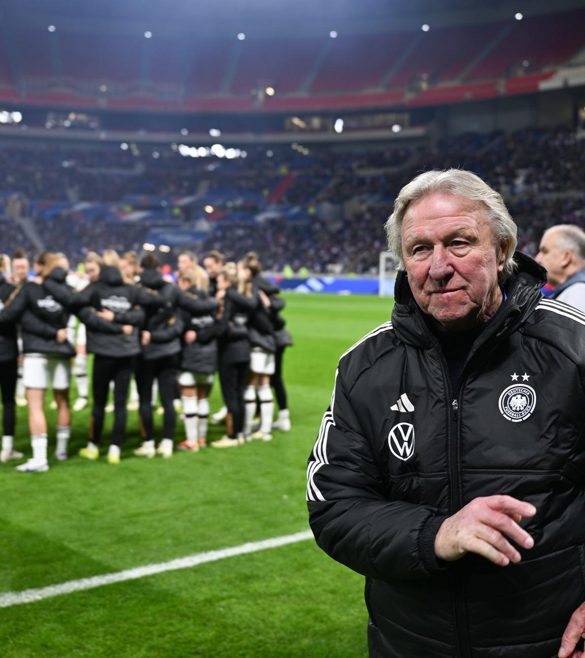 Im Vordergrund steht Horst Hrubesch. Im Hintergrund steht die Fußballnationalmannschaft der Frauen im Kreis.