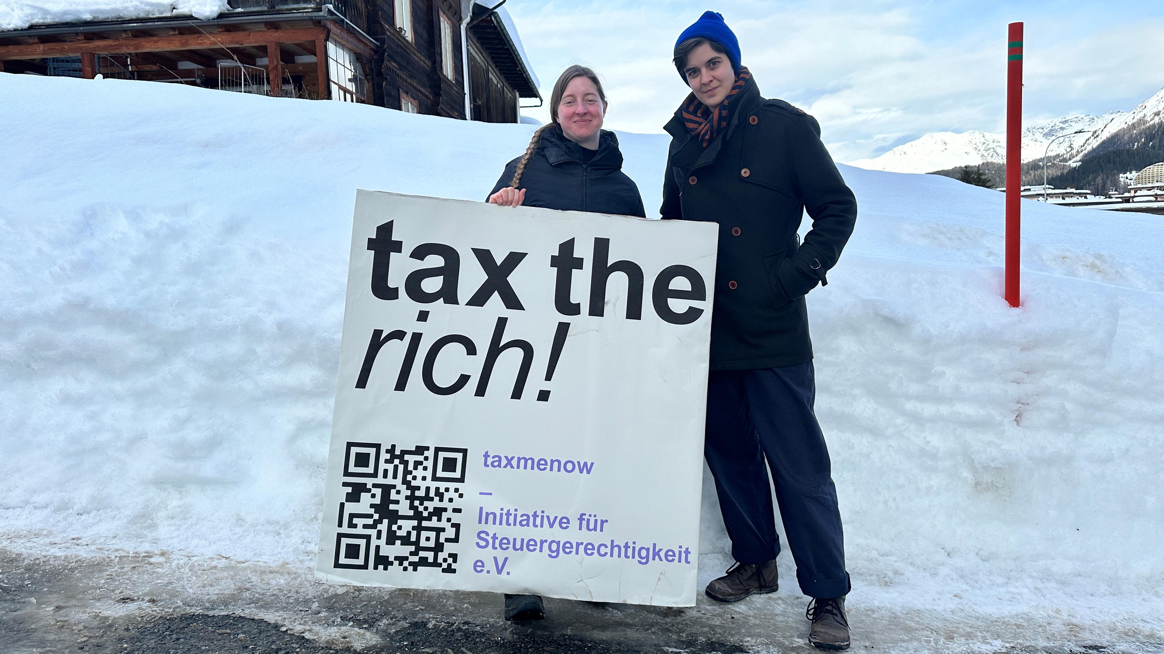 Zwei Millionenerbinnen, Stefanie Bremer (l.) und Marlene Engelhorn (r.), halten ein Schild mit der Aufschrift "Tax the rich". Sie stehen in einer Winterlandschaft und tragen warme Kleidung.