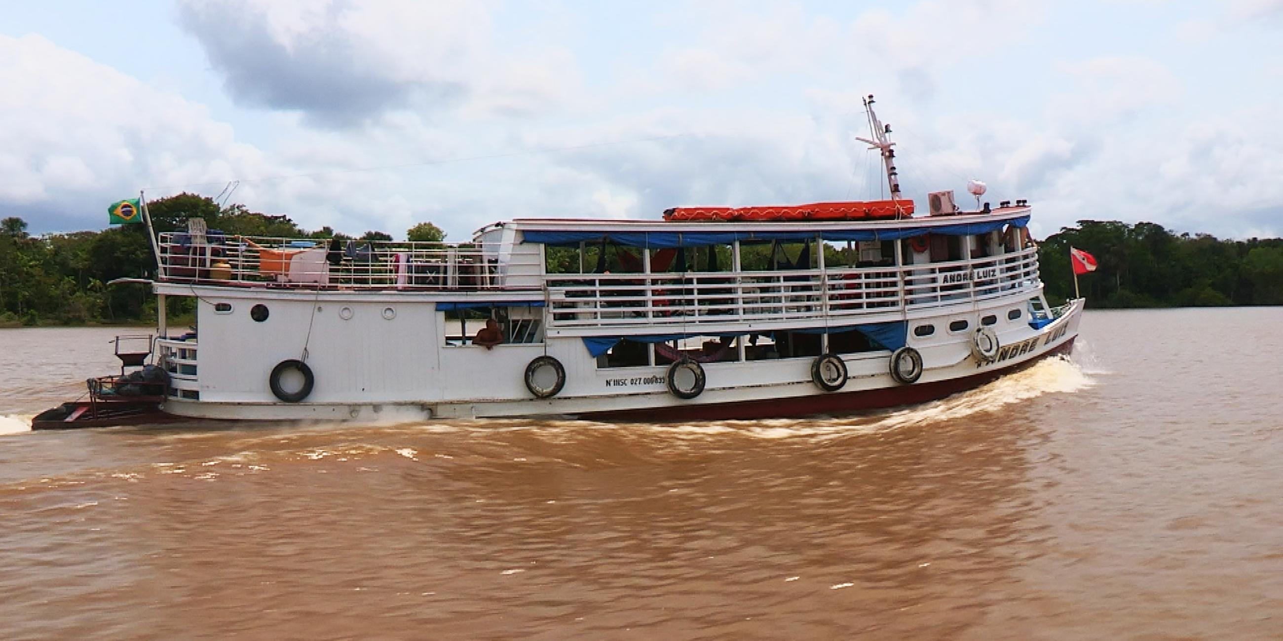  Auf einem bräunlichen Gewässer mit leichten Wellen fährt ein weißes, einstöckiges Schiff. Auf dem Dach des Schiffes liegen große, flache rote Gegenstände, mutmaßlich Schlauchboote. Im Hintergrund sind ein blauer, mit großen Wolken durchsetzter Himmel sowie dichte grüne Bäume zu sehen.